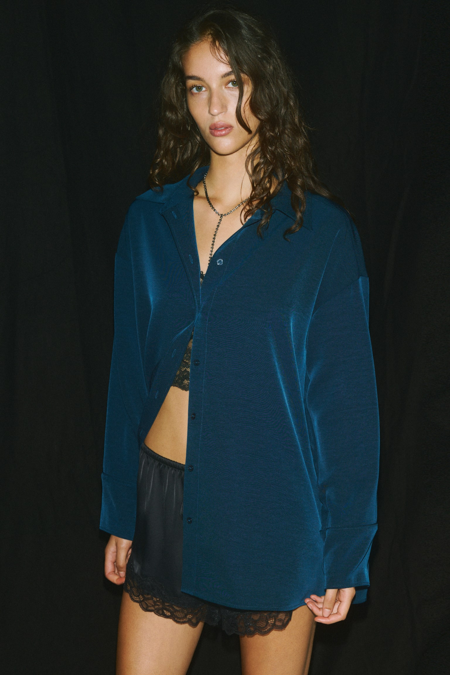 An oversized dark teal shirt is hanging facing the front, displaying a pointed collar, a button-down front, and dropped shoulders. The design is featuring long sleeves with wide, double-layered cuffs and a straight-cut hem in a softly draping, textured fabric. - 1