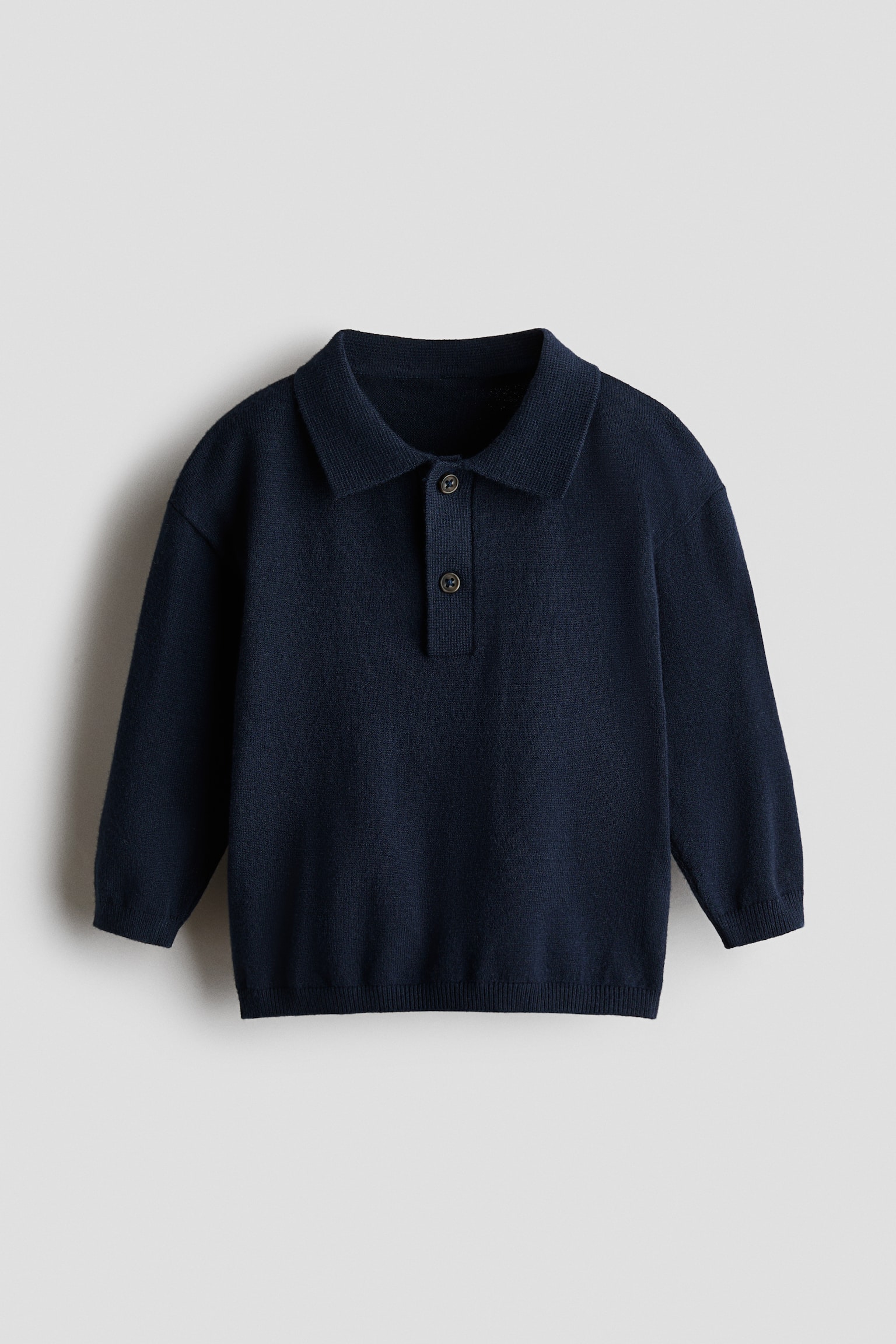 Een marineblauwe, fijngebreide rugbyshirt met lange mouwen wordt van voren getoond, met een klassieke kraag, een knoopsluiting met twee knopen en verlaagde schoudernaden. Het shirt heeft geribbelde details bij de manchetten en de zoom.