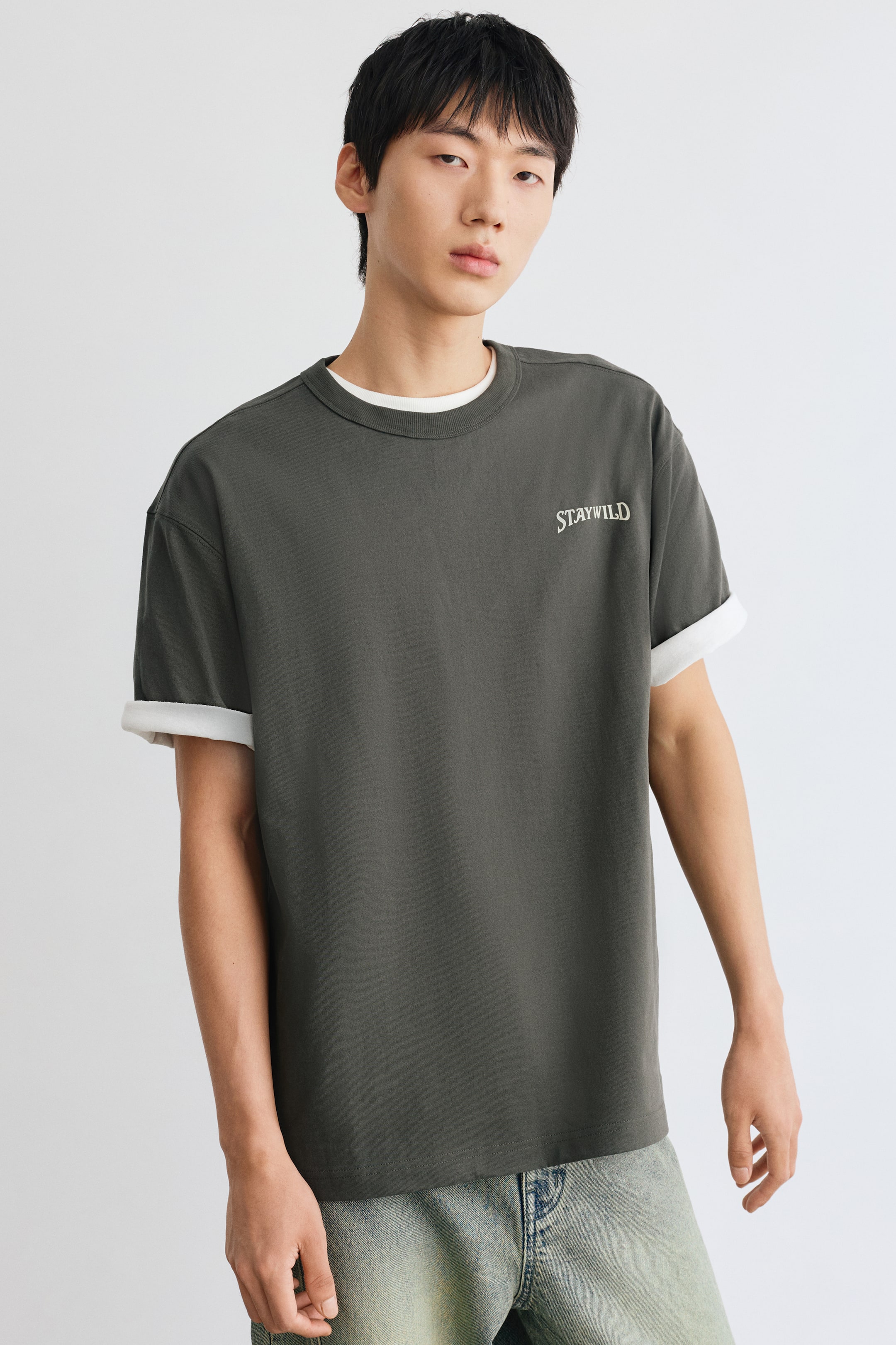 View larger image: A dark grey, loose-fit T-shirt featuring a small white "STAYWILD" print on the left chest is worn by the model. The T-shirt is layered over a white tee, which is visible at the round, ribbed neckline and where the sleeves are rolled up.