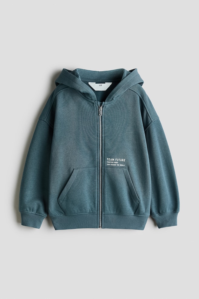 Eine Vorderansicht eines tealblauen Zip-up-Hoodies zeigt eine angesetzte Kapuze, einen durchgehenden FrontreiÃverschluss und eine geteilte KÃ¤ngurutasche. Der Hoodie verfÃ¼gt Ã¼ber Ã¼berschnittene Schultern, gerippte BÃ¼ndchen, einen gerippten Saum und kleinen weiÃen Aufdruck auf der linken Brustseite. - 1