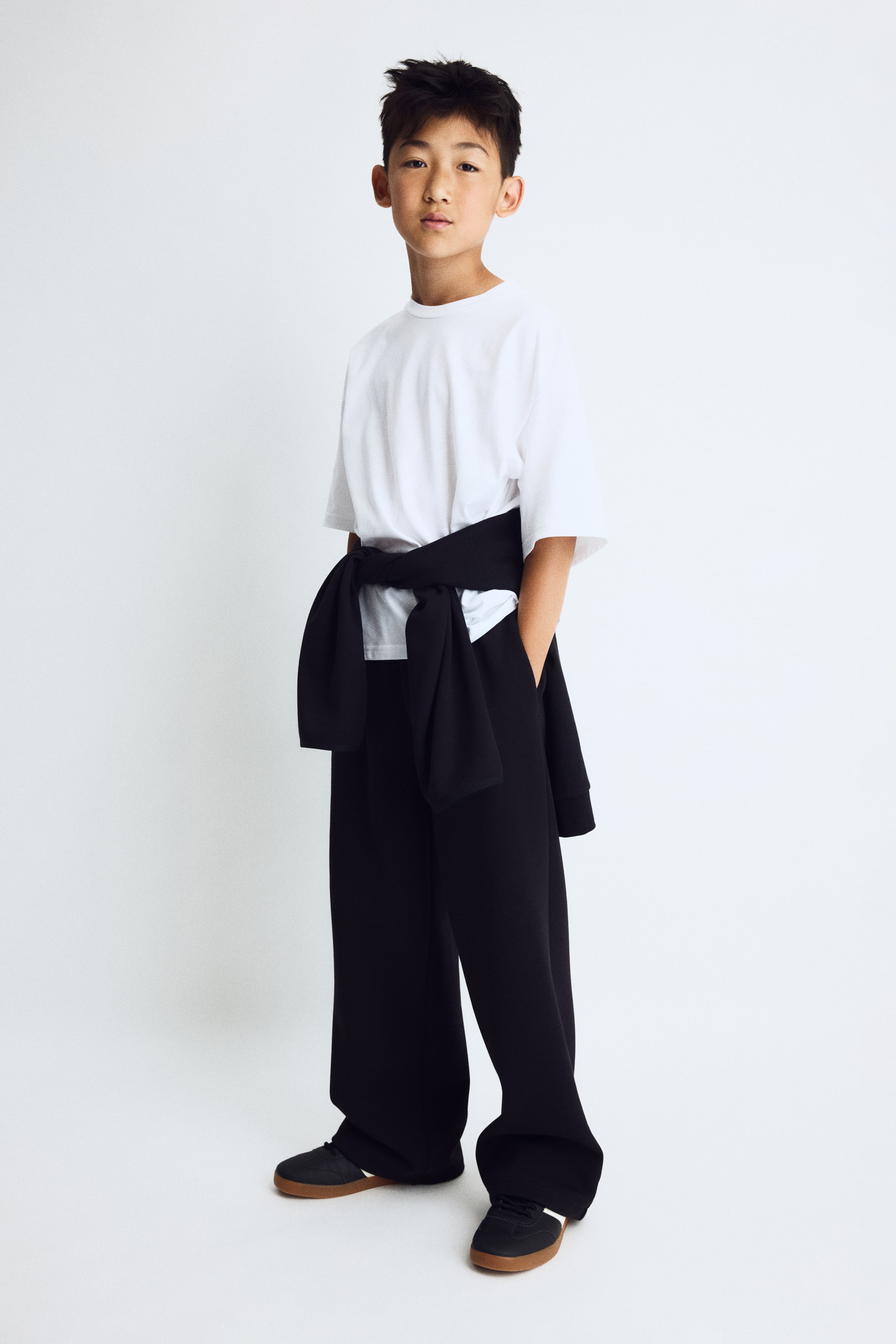 Voir une image plus grande: Les pantalons de jogging à coupe ample sont présentés de face en noir, avec une coupe à jambes larges et des poches latérales, une main reposant dans une poche. Un chandail noir est noué autour de la taille, cachant la ceinture.