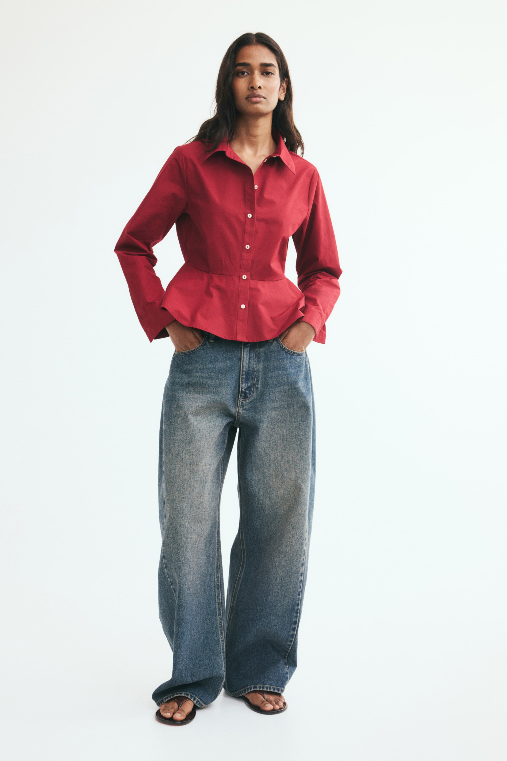 View larger image: A red cotton poplin shirt featuring a collar, front buttons, and a flared peplum hem is being worn by the model. The shirt is styled with wide-leg blue jeans, with the model placing hands in the pockets.
