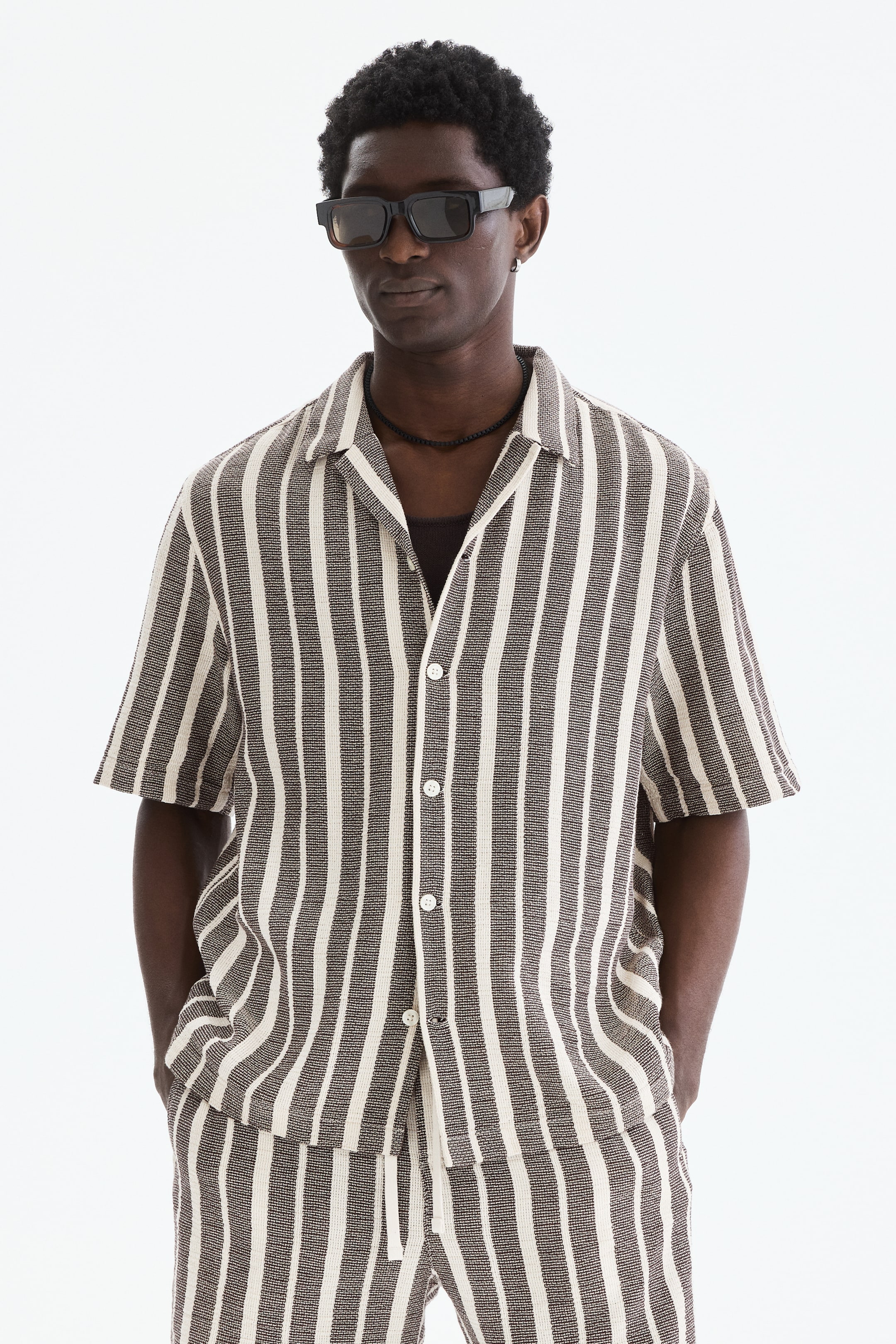 View larger image: A striped short-sleeve shirt and matching shorts are displayed from a front-facing angle. The shirt features an open collar and button-front closure, revealing a dark undershirt. Both pieces showcase vertical dark brown and off-white stripes with a visible woven texture, and the shorts include a drawstring waist.