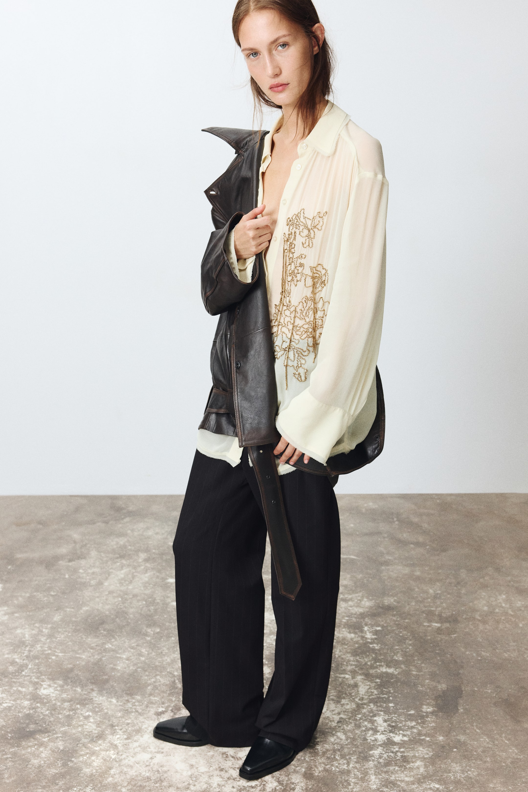View larger image: Loose-fit tailored pants in black twill are worn by the model, featuring a straight-leg silhouette with visible creases down the front. The pants are styled with a sheer embroidered cream shirt and black leather shoes, while a dark brown leather jacket is draped over one shoulder.