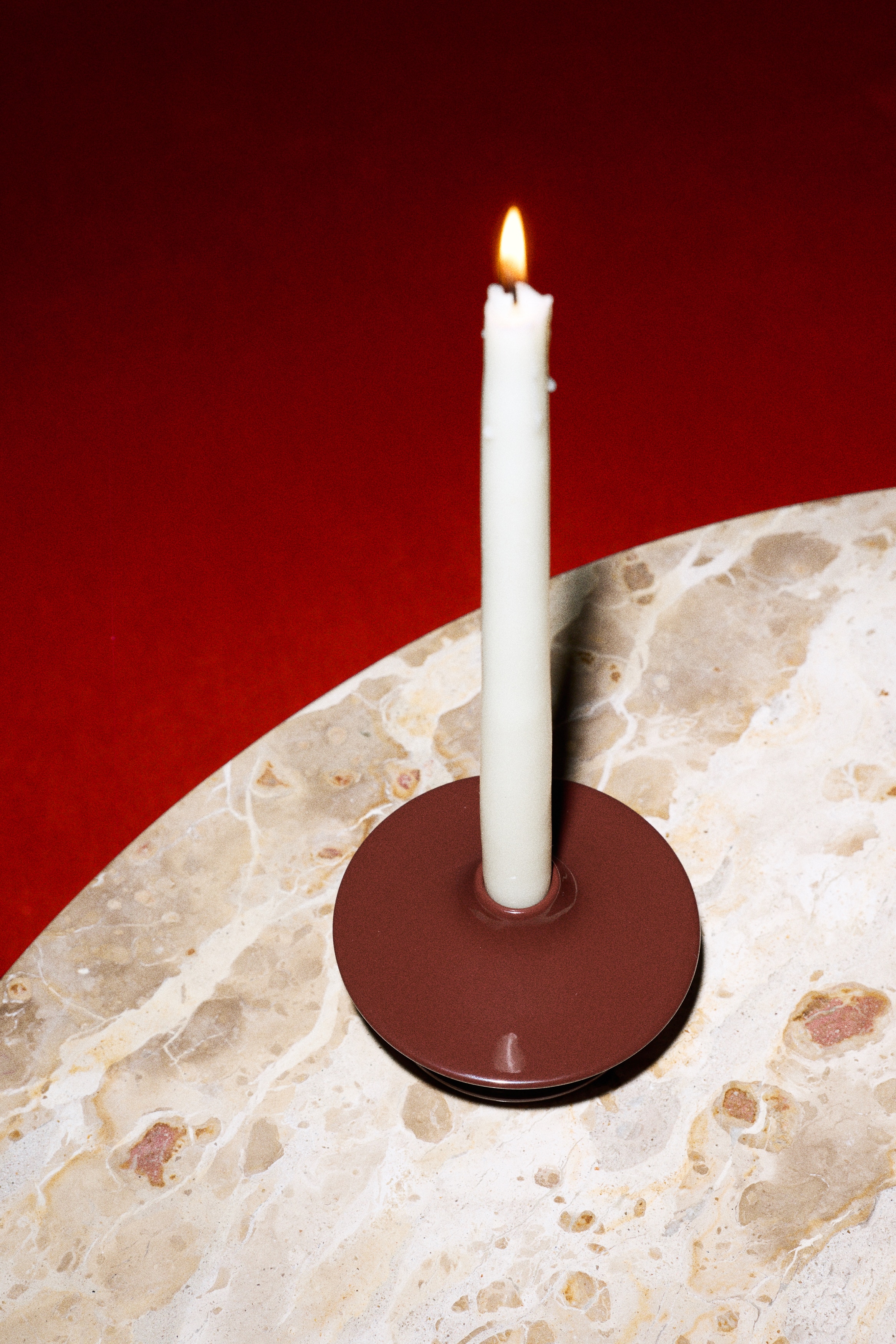 A white taper candle is standing upright in a dark red, glossy candle holder. The holder is designed with a two-tiered, sculptural form, featuring a wide top disc and a smaller base disc.