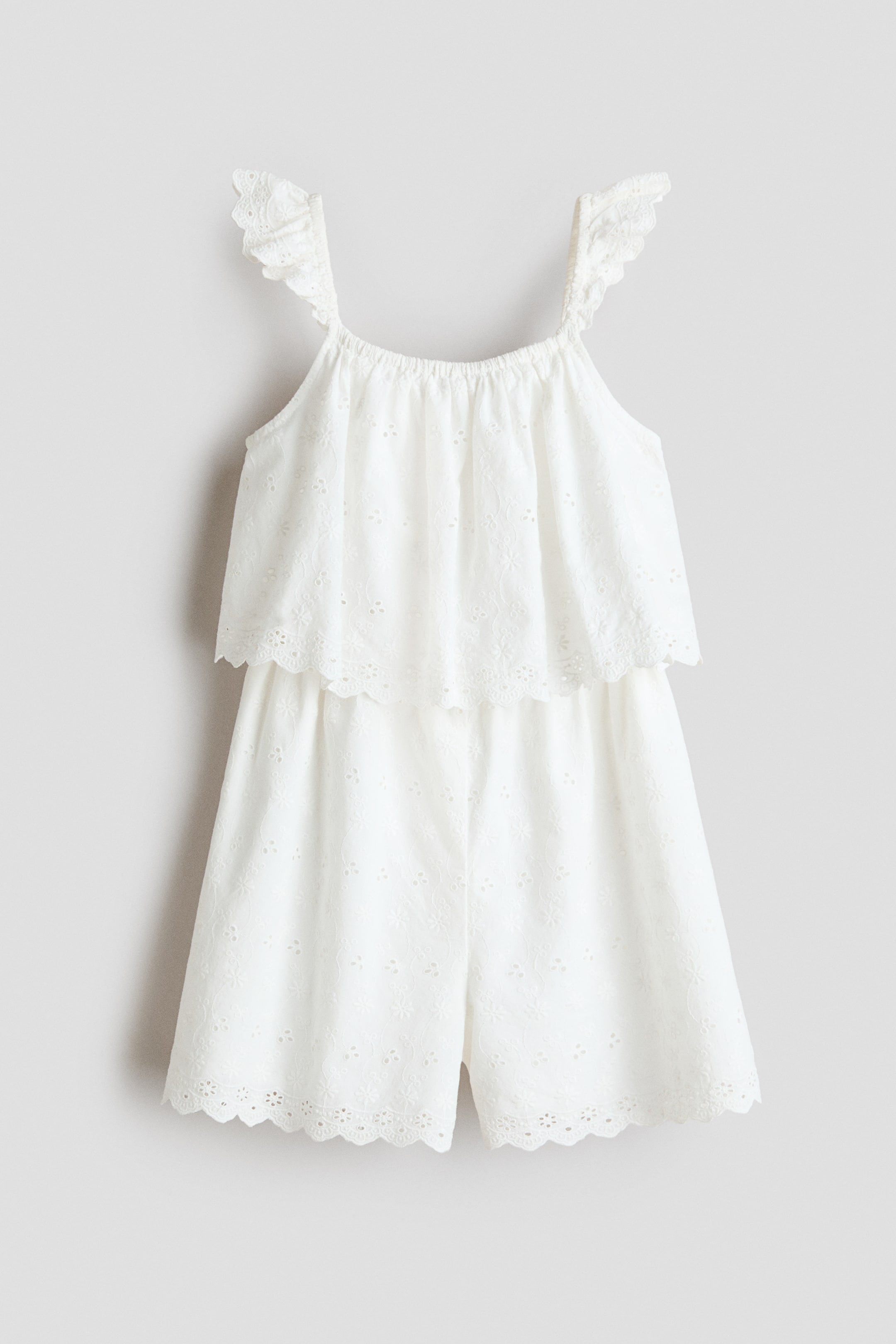 Grotere afbeelding bekijken: Een witte playsuit van ajourkant wordt frontaal gepresenteerd, met dunne ruchesbandjes en een elastische, geplooide halslijn. Het lijfje toont een gelaagd ontwerp met een geschulpte, geborduurde rand, en de shorts hebben een bijpassende geschulpte zoom.
