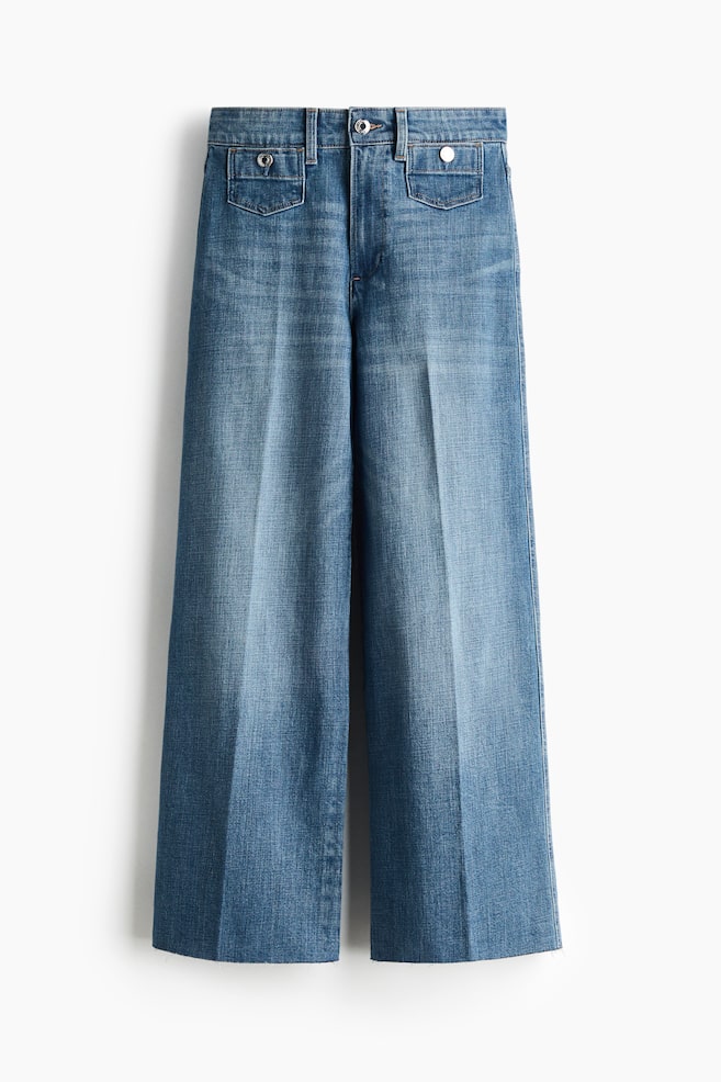 Blue denim jeans are facing FRONT, featuring a high waist with a zip fly and a single button closure. Two patch front pockets with button details are visible at the waist, and creases are running down the front of each wide leg, which is presenting a slim fit from the waist to the hip and a wider cut from the thigh to the ankle-length hem. - 1