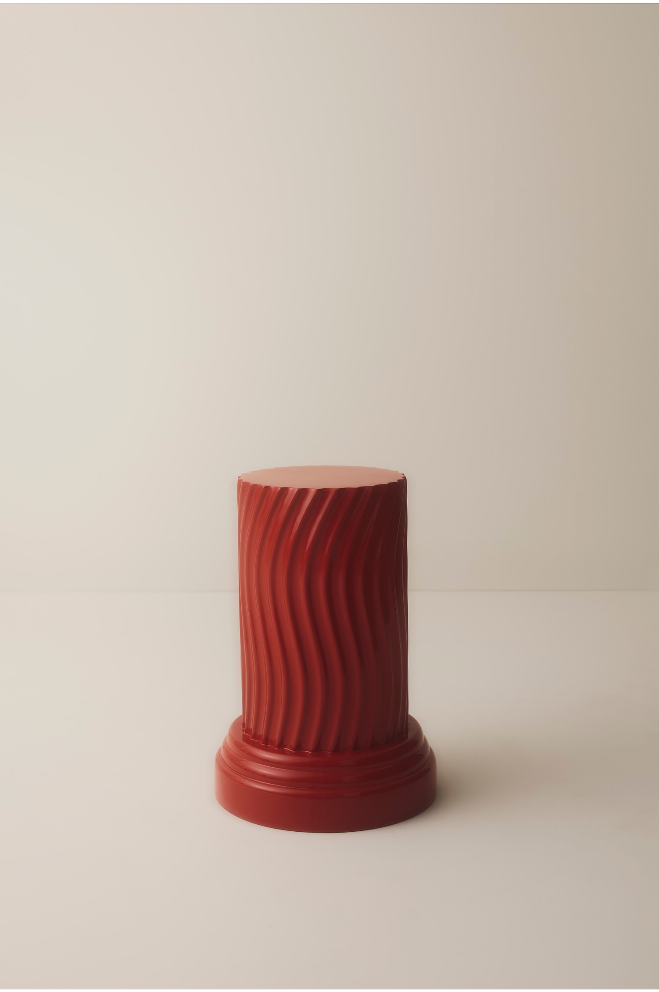 Visualizza immagine più grande: Un piedistallo rosso lucido in stile colonna è in posizione eretta, con un corpo cilindrico caratterizzato da scanalature verticali ondulate e da un piano superiore circolare e piatto. Il piedistallo poggia su una base circolare più ampia e a gradini.