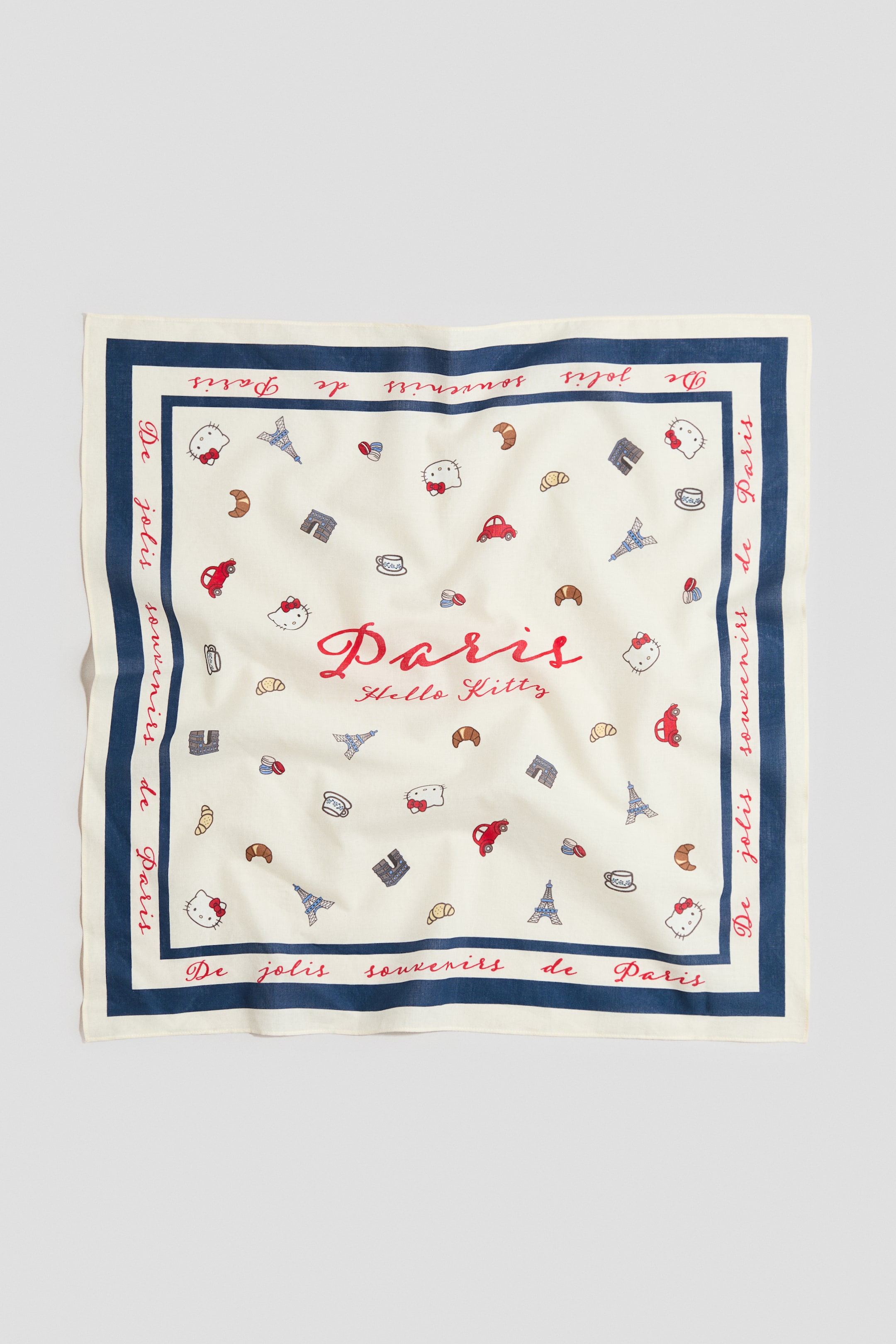 Größeres Bild ansehen: Ein quadratisches Tuch aus einem luftigen, bedruckten Baumwollgewebe liegt flach da und präsentiert eine cremefarbene Basis mit einem marineblauen Rand. Der Rand zeigt den Text "De jolis souvenirs de Paris", während das Zentrum den Text "Paris Hello Kitty" in Rot darstellt, umgeben von verstreuten Motiven von Hello-Kitty-Köpfen, Eiffeltürmen, Croissants und anderen Pariser Elementen.