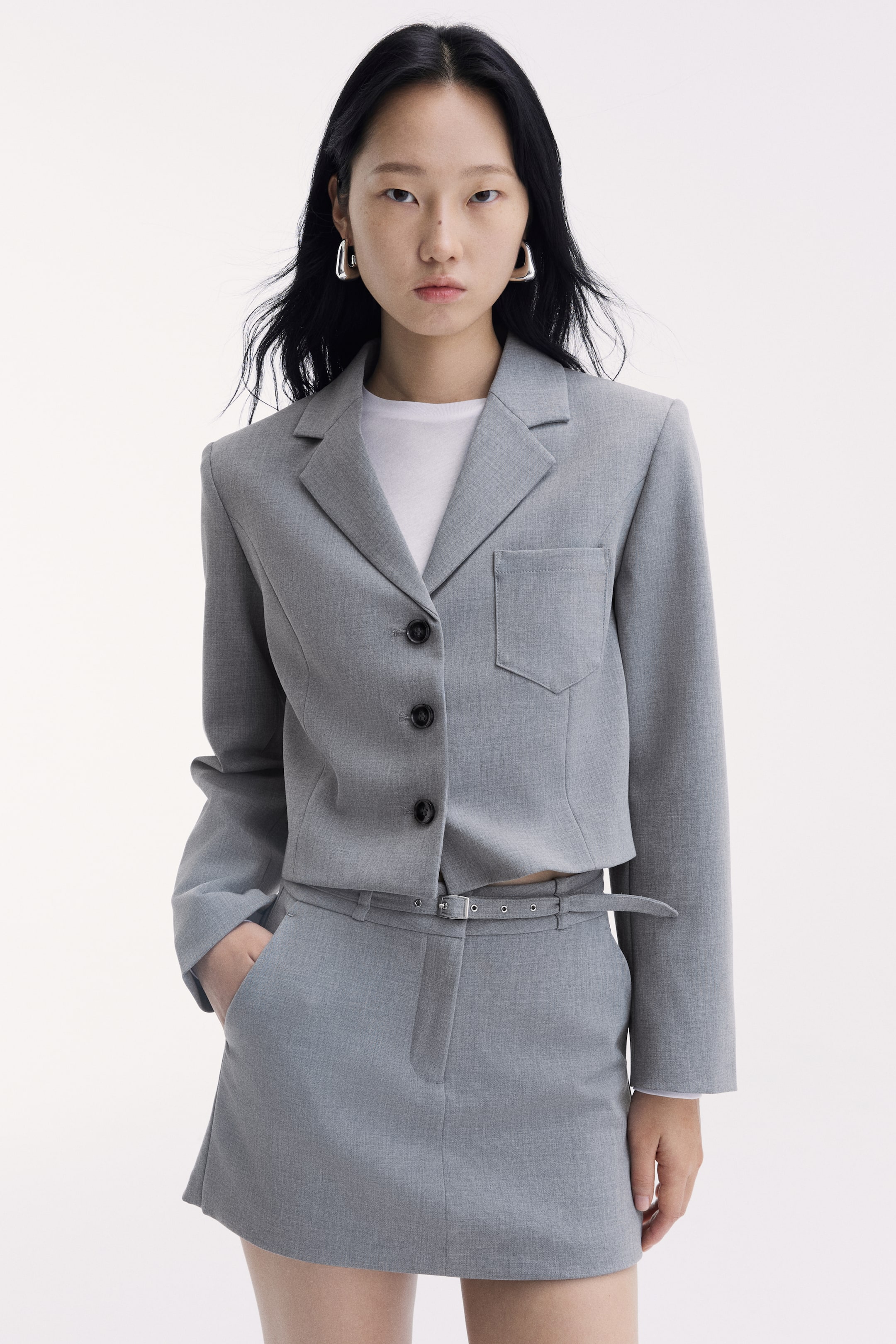 View larger image: The model is wearing a short grey skirt with a regular waist and a narrow adjustable belt with a metal buckle, with one hand resting in a diagonal side pocket. The skirt is styled with a matching cropped grey blazer layered over a white T-shirt.