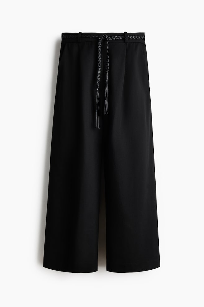 Pantalones negros de pierna ancha se muestran desde el frente, con una cinturilla plana con trabillas y un cinturón negro trenzado y delgado pasado por las trabillas, atado en el centro con dos borlas colgantes. Los pantalones están diseñados con un corte relajado y de pierna ancha, terminando por encima del tobillo. - 1
