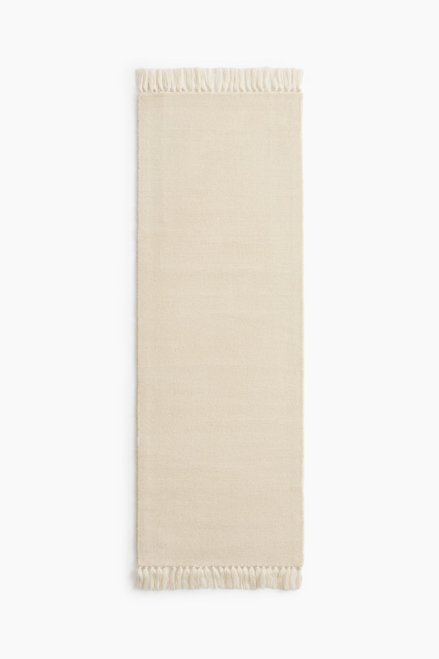 A cream-colored runner rug is shown from a flat overhead view, displaying a solid-colored textured body with subtle horizontal ribbing. Thick, knotted tasseled fringes are trimming both of the shorter ends.