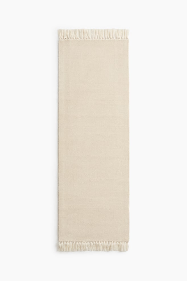 Un tapis de couloir de couleur crème est présenté en vue plongeante à plat, mettant en valeur un corps texturé uni avec de subtiles côtes horizontales. De grosses franges à pompons noués ornent les deux extrémités les plus courtes. - 1