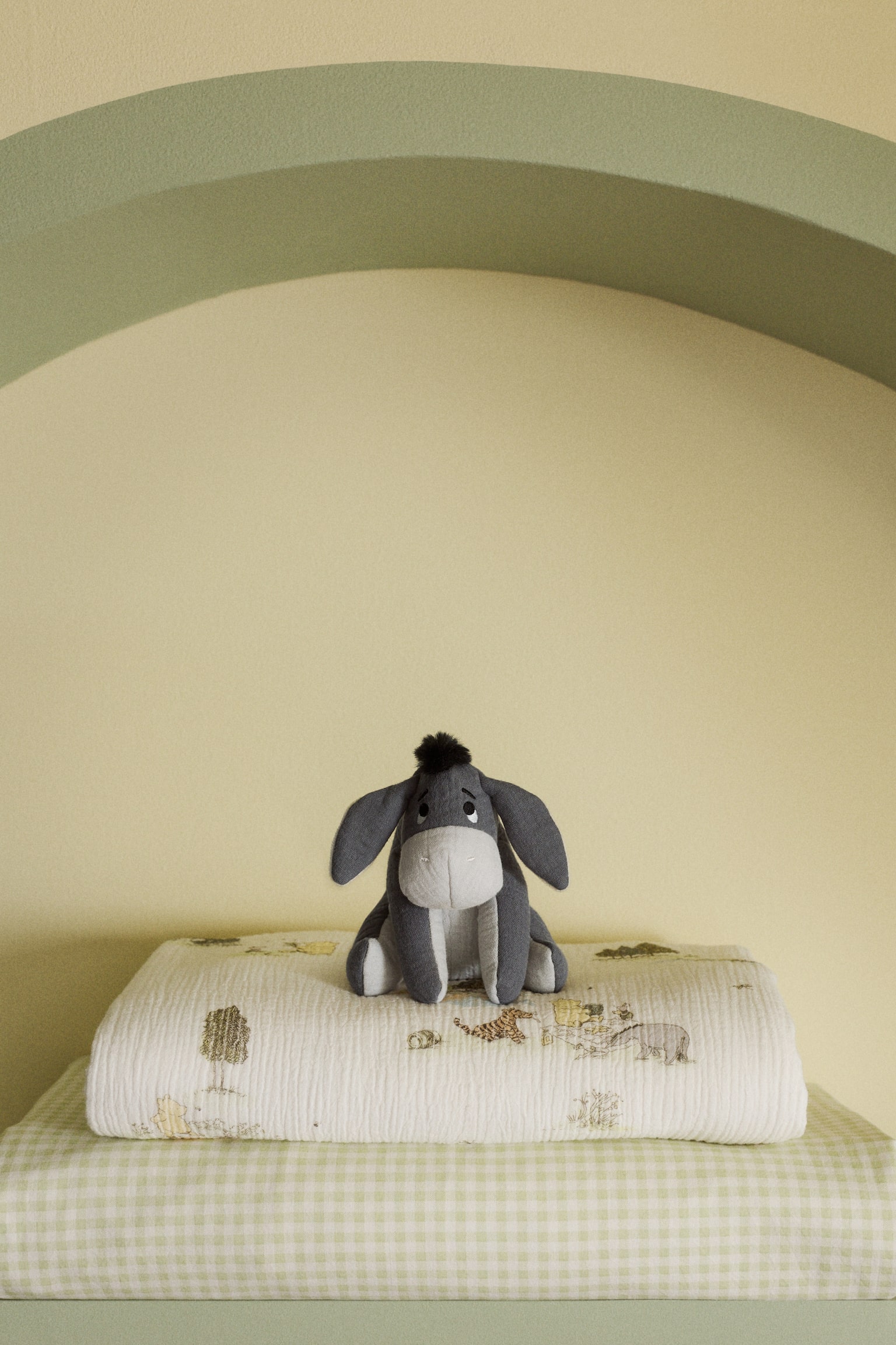 A grey Eeyore plush toy is sitting facing forward on a cream-coloured muslin blanket. The blanket features illustrations of animals and trees and is layered over a light green and white gingham blanket. - 1