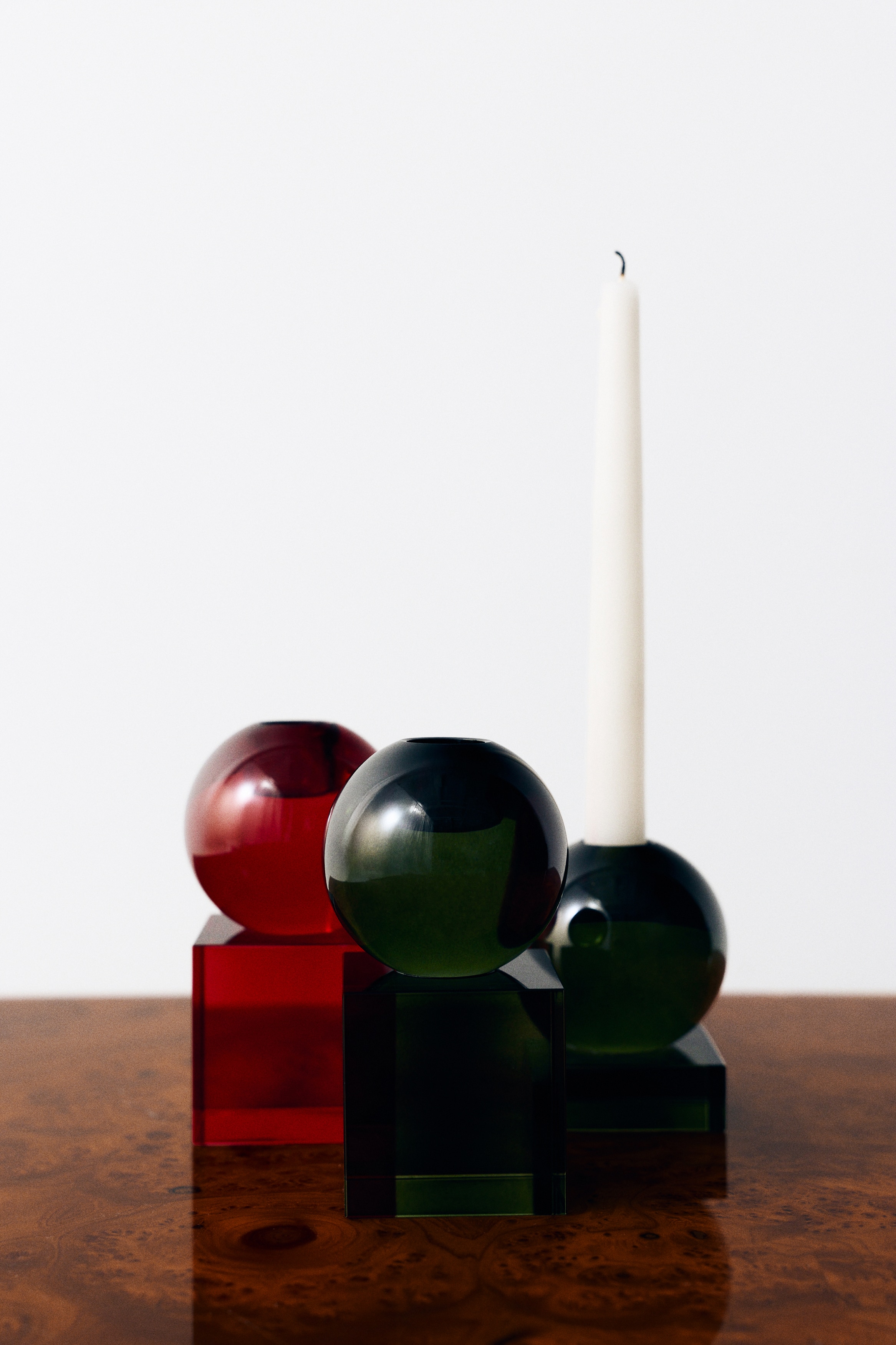 Un portacandela in vetro verde, mostrato da un angolo frontale, sostiene una candela alta e bianca a candela lunga. Il portacandela presenta un design geometrico moderno con una base a cubo in vetro verde oliva traslucido sormontata da una sfera in vetro coordinata.