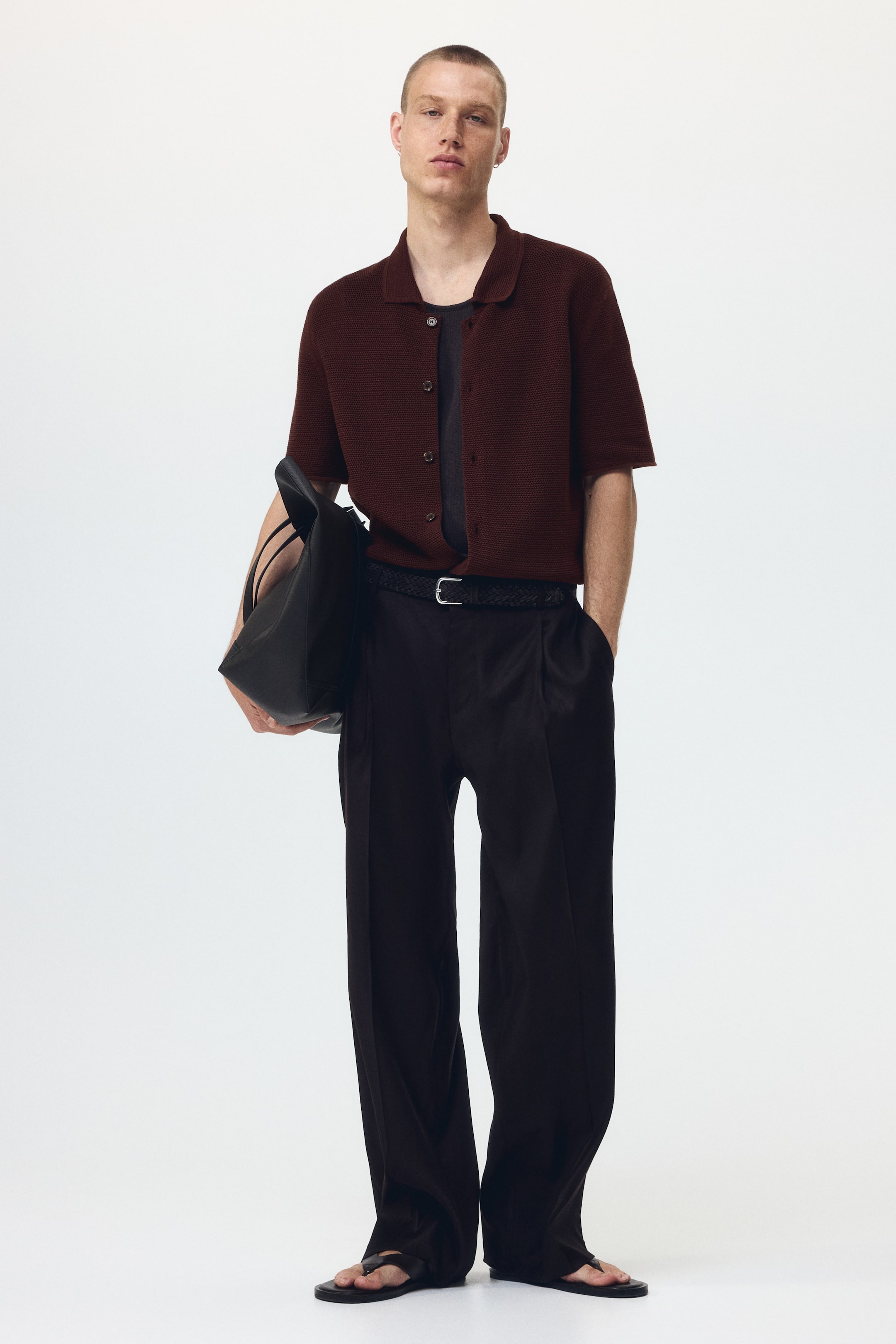 Ver imagen más grande: Una camiseta polo de punto de manga corta en color granate se combina con una camiseta de manga corta en gris oscuro, estilizada con pantalones plisados de pierna ancha en negro, un cinturón trenzado negro y sandalias de dedo negras. Se sostiene un bolso tote negro.