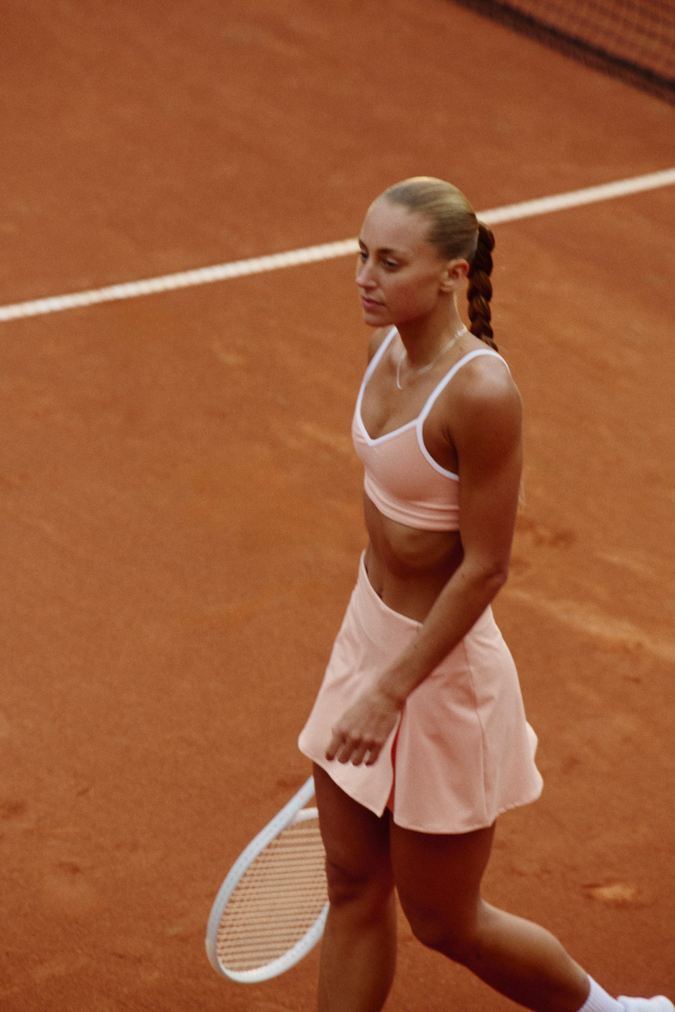 Ingrandisci l'immagine: Un completo sportivo color pesca chiaro è indossato, composto da un reggiseno sportivo a V con rifiniture bianche e spalline sottili, e una gonna da tennis svasata abbinata con un sottile design a portafoglio sul davanti. Una racchetta da tennis bianca è tenuta accanto alla gonna.