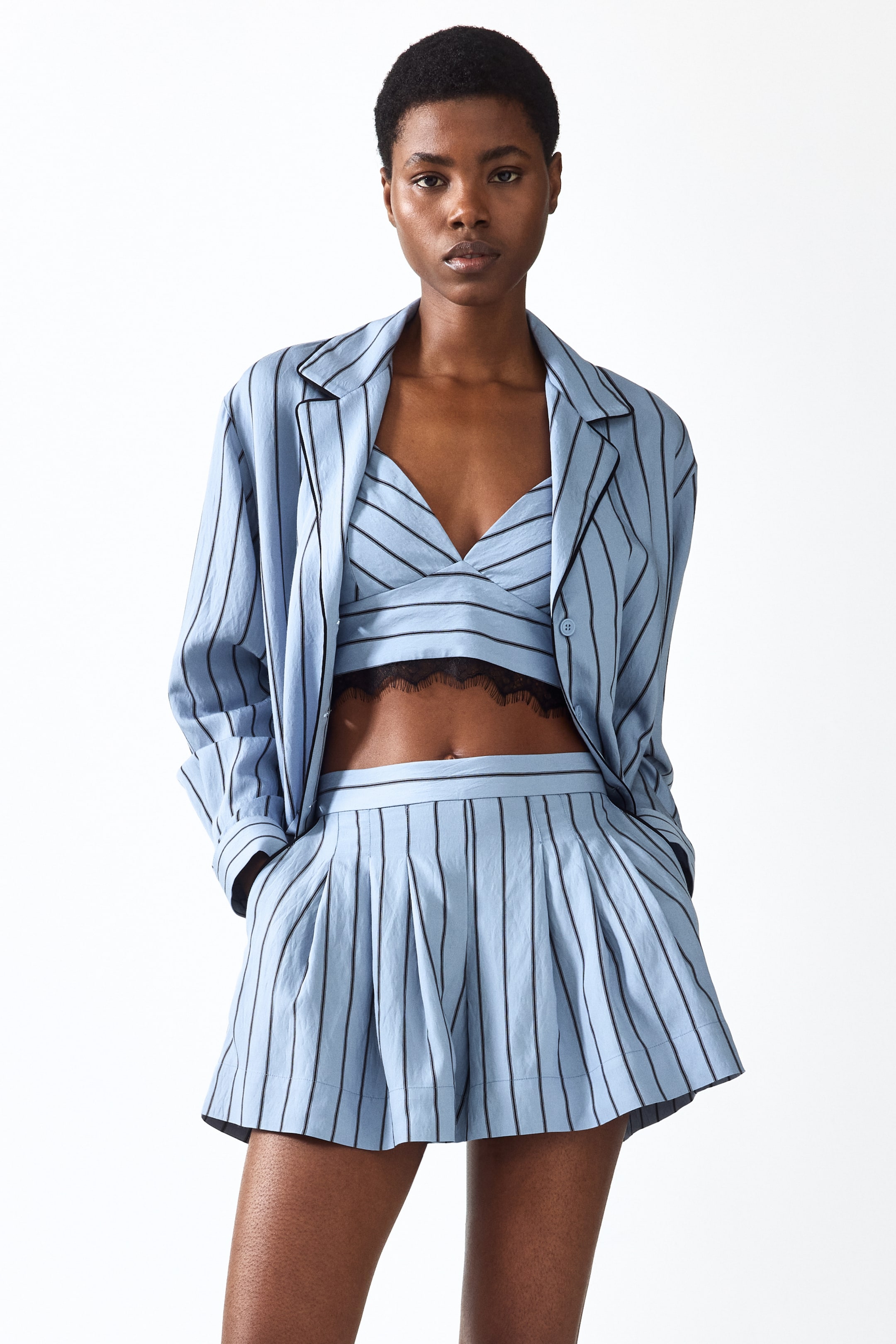 Ingrandisci l'immagine: La modella indossa pantaloncini a righe azzurro chiaro con vestibilità ampia. I pantaloncini presentano una vita alta, pieghe frontali e tasche laterali diagonali con bordature nere, nelle quali la modella inserisce le mani.