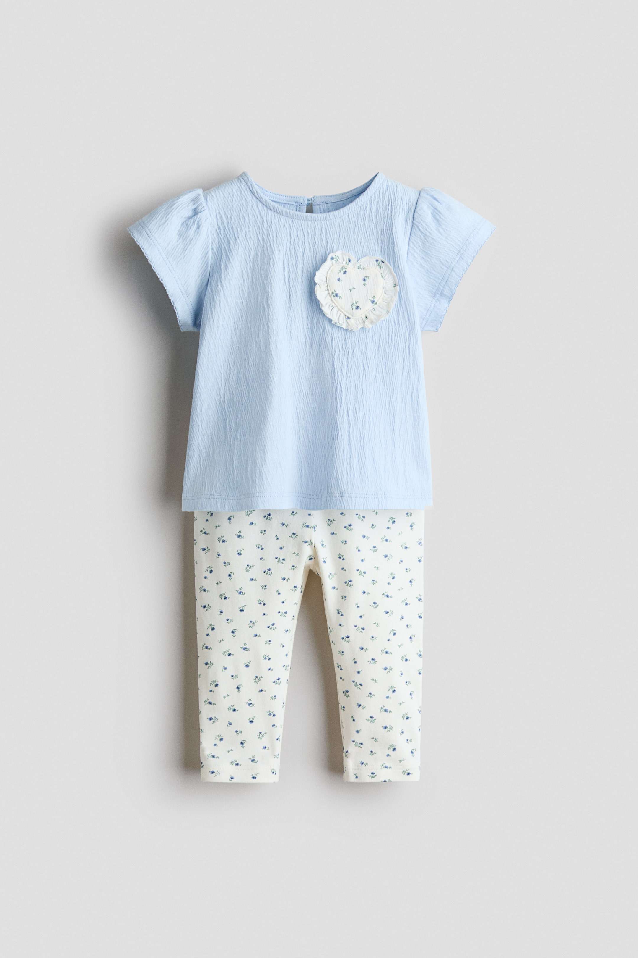 View larger image: A two-piece baby outfit is presented, facing front, with a light blue textured top and cream-colored pants. The top features short puffed sleeves, a round neckline, and a circular appliqué showcasing a white ruffled edge and a small blue floral print, while the pants display an all-over pattern of tiny blue flowers.