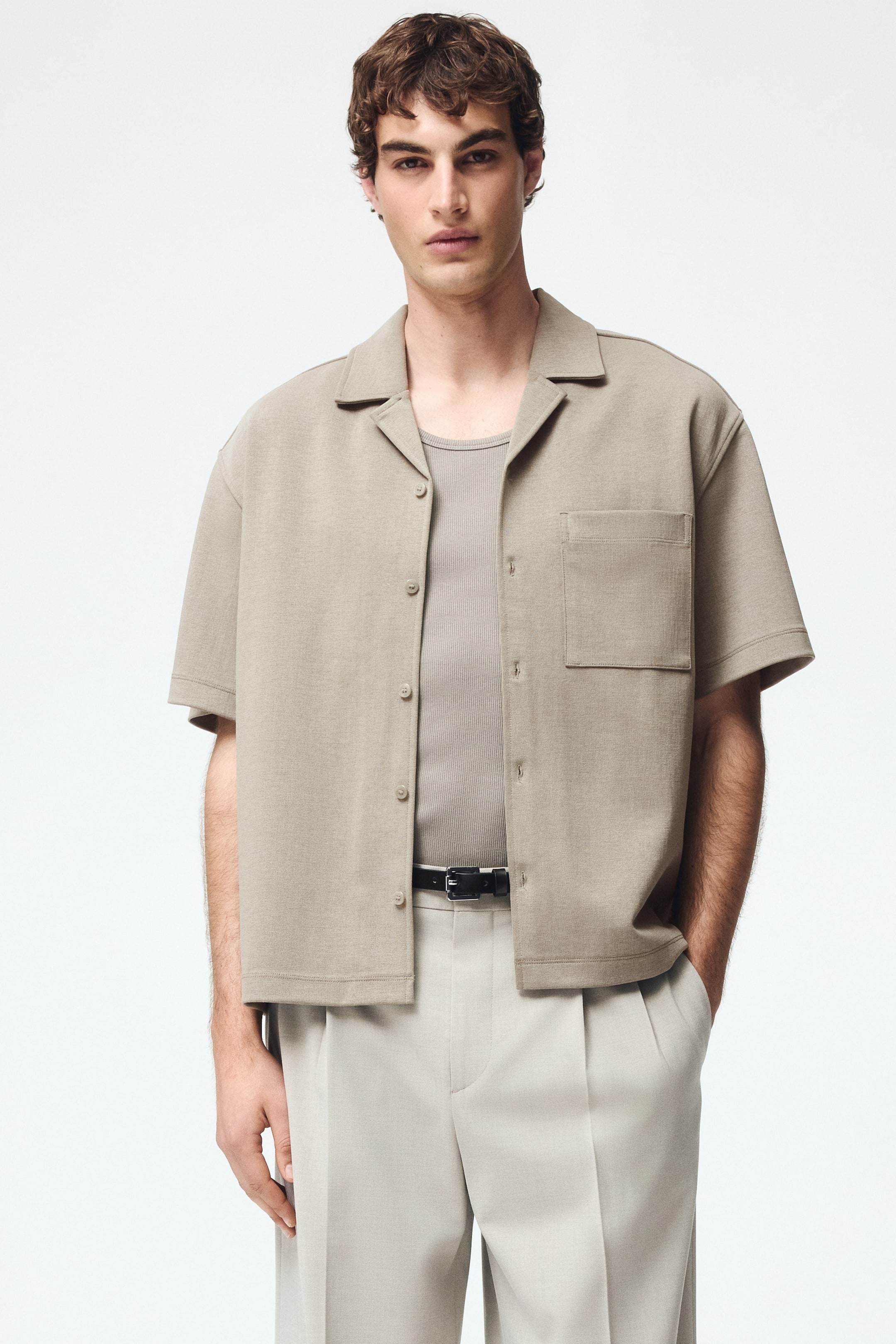 Ver imagen más grande: Una camisa de punto de manga corta de color marrón claro presenta un cuello tipo campamento, mangas cortas y un bolsillo de parche único en el pecho izquierdo, con botones que se extienden por la parte delantera. Se lleva abierta sobre una camiseta de tirantes acanalada.