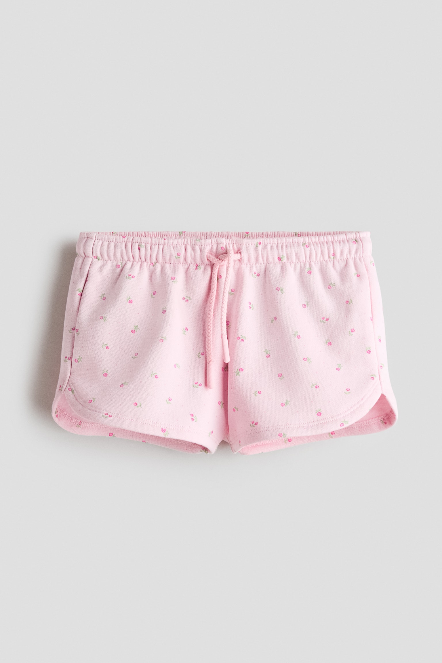 En frontvy av ljusrosa sweatshorts som visar ett heltÃ¤ckande tryck av smÃ¥ rosa blomknoppar och grÃ¶na blad. Shortsen har en elastisk midja med matchande dragsko, diskreta sidofickor och rundade, svÃ¤ngda fÃ¥llar.