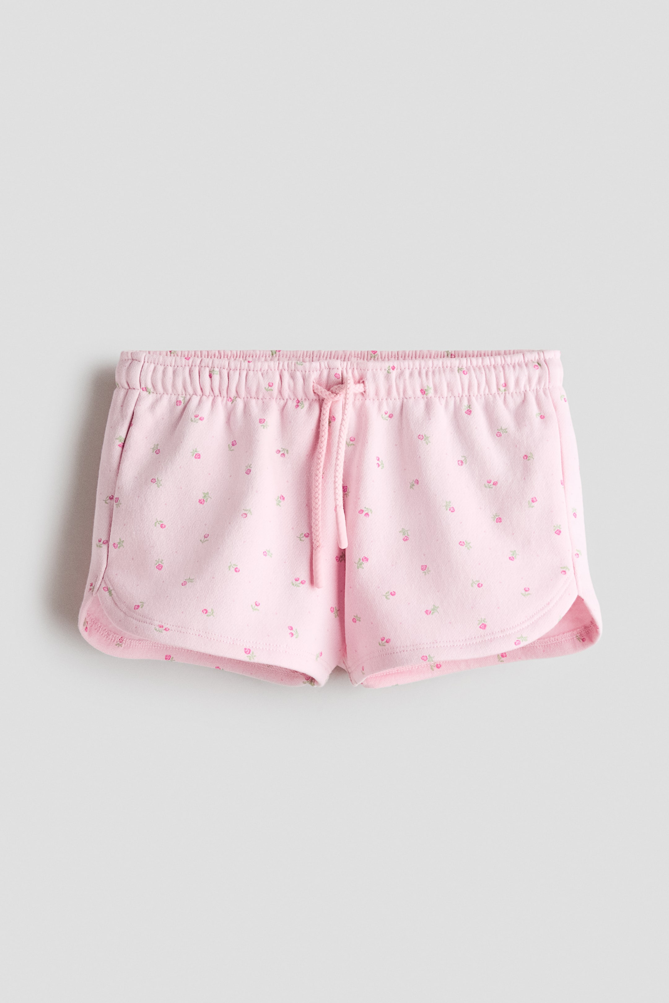 Visa större bild: En framifrånvy av ljusrosa sweatshorts visar ett heltryck med små rosa blomknoppar och gröna blad. Shortsen har en resårmidja med matchande dragsko, diskreta sidfickor och rundade, böjda fållar.