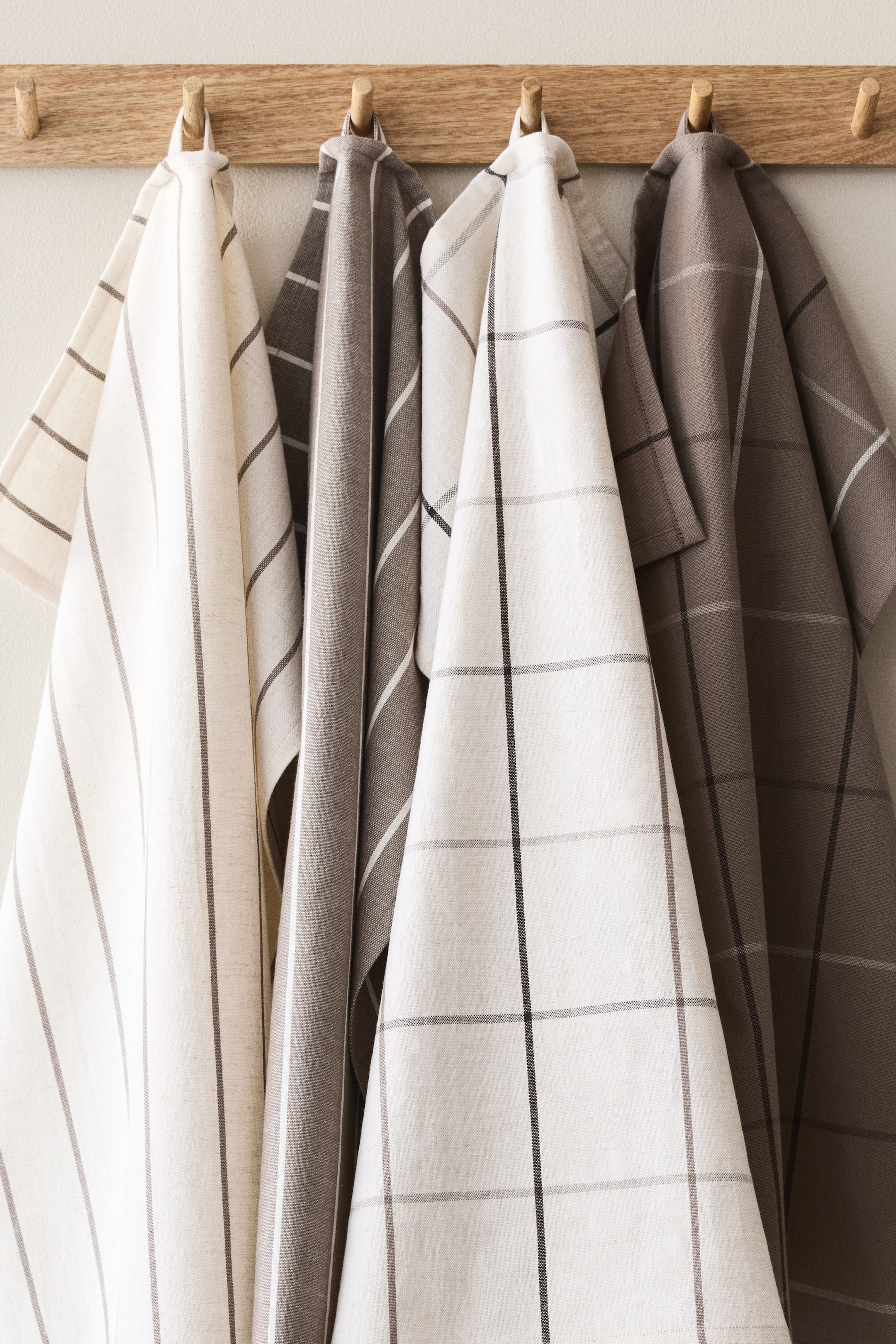 View larger image: Four kitchen towels hang vertically from wooden pegs. These towels showcase striped and plaid patterns in off-white and taupe tones, highlighted by contrasting dark and light lines. Each towel displays a distinct woven texture.