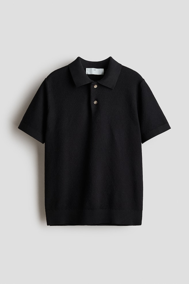 Ein schwarzes Poloshirt zeigt seine Vorderansicht mit einer Waffelstrickstruktur. Es ist mit einem Kragen, einer Zwei-Knopf-Leiste, überschnittenen Schultern und kurzen Ärmeln gestaltet und wird mit gerippten Bündchen und einem gerippten Saum abgeschlossen. - 1