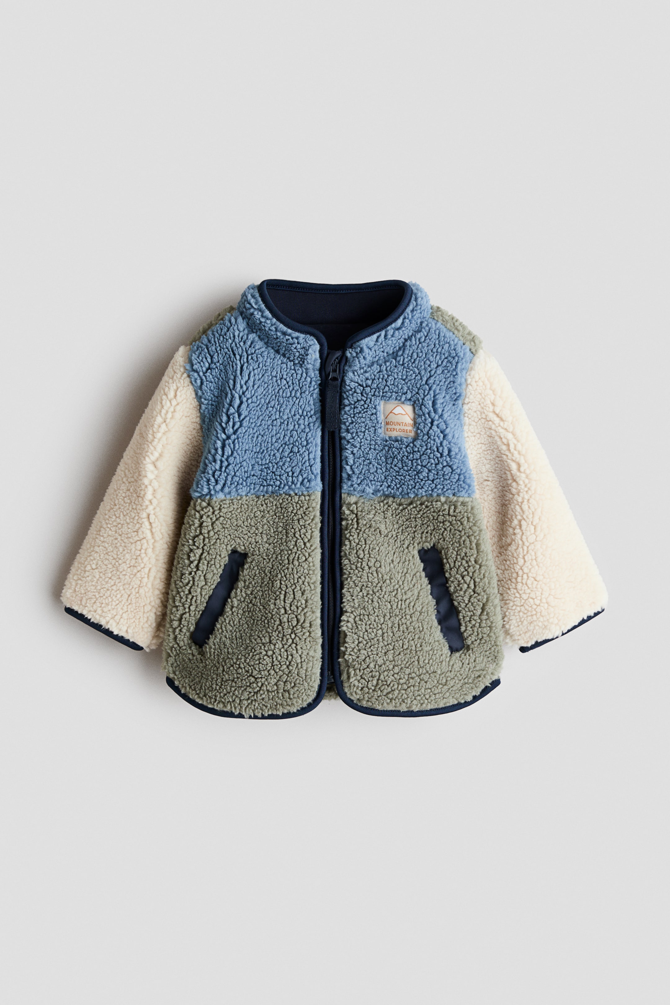 View larger image: A children's front-facing sherpa fleece jacket showcases a colour-blocked design with blue on the upper body, sage green on the lower body, and cream on the sleeves. It features a dark navy stand-up collar, a full-length zip closure, dark navy binding along the hem, cuffs, and two front welt pockets, and a small "Mountain Explorer" patch on the right chest.