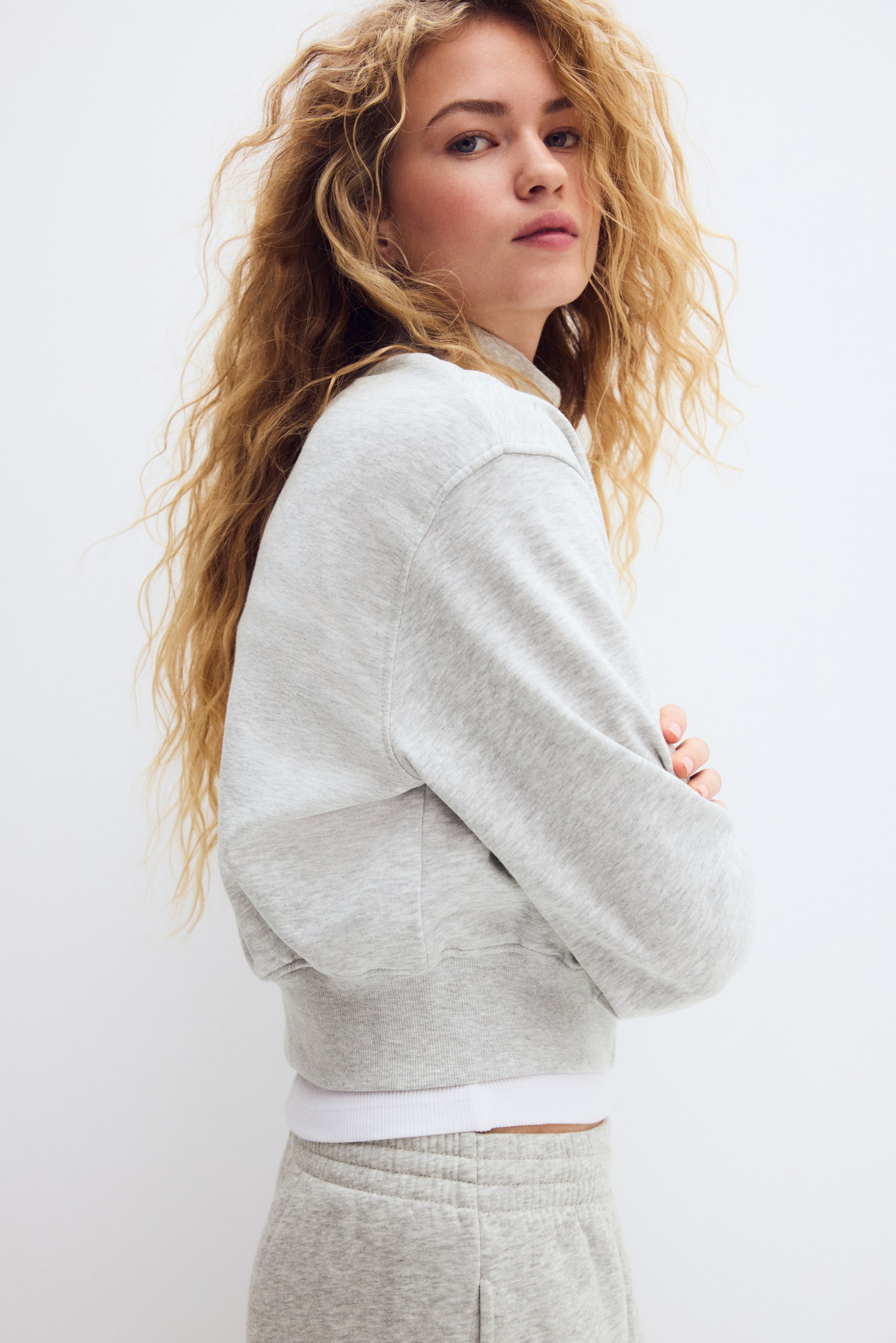 View larger image: A light grey cropped sweatshirt features a mock neck, ribbed cuffs and a ribbed hem, shown from a side and slightly rear angle. It is layered over a white ribbed band, with matching light grey sweatpants visible below.