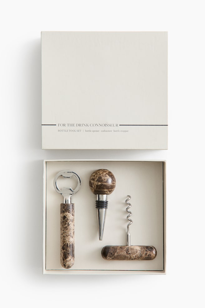 A bottle tool set is presented in an open, light beige gift box, with its lid reading 'FOR THE DRINK CONNOISSEUR' and listing the contents as 'BOTTLE TOOL SET \| bottle opener corkscrew bottle stopper'. Inside, a bottle opener, a spherical-topped bottle stopper, and a compact corkscrew are securely held by white elastic bands, all featuring brown and beige marbled handles. - 1