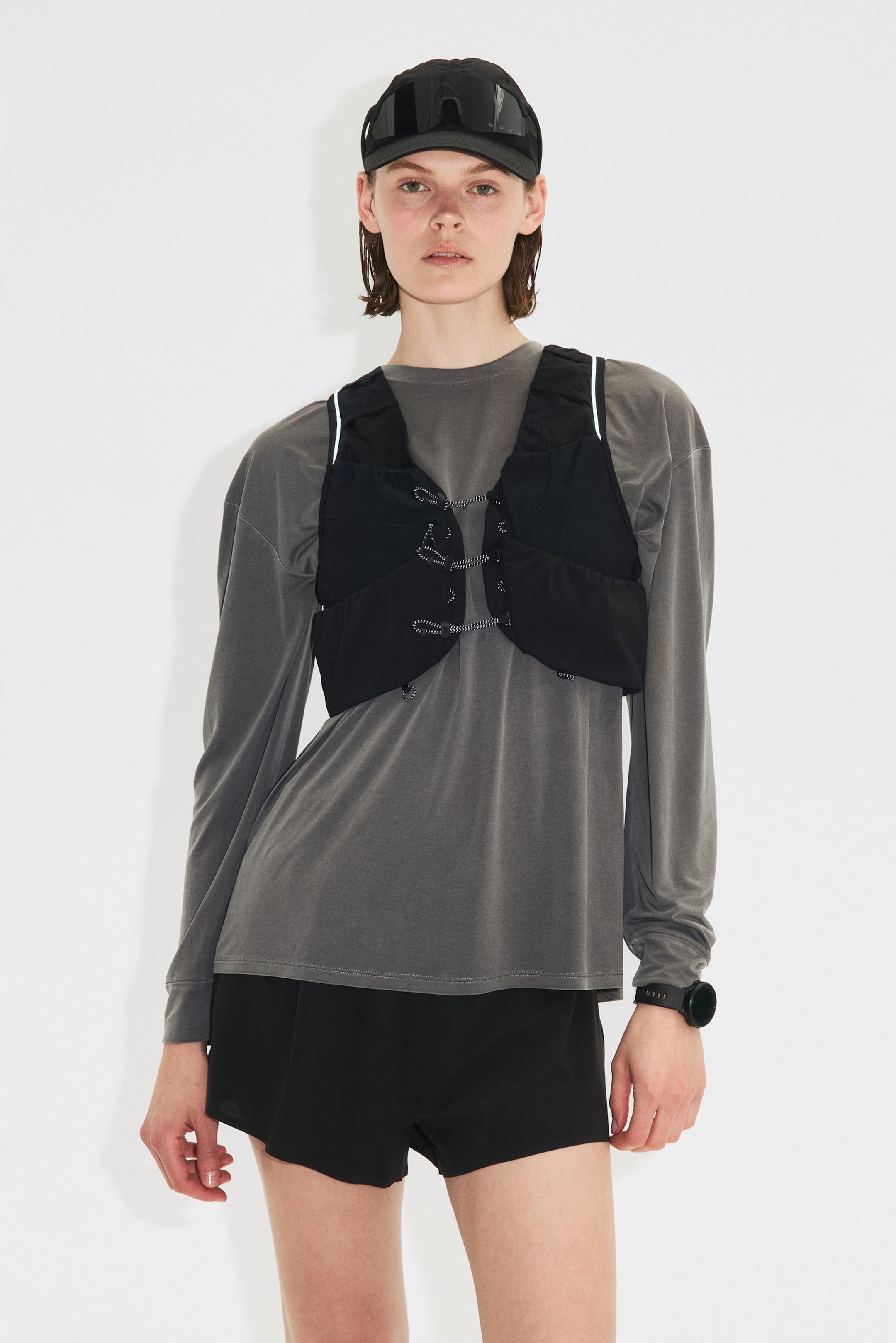 Grotere afbeelding bekijken: Een zwarte, cropped technische bodywarmer wordt gepresenteerd onder een lichte hoek, met een V-hals en drie sets witte koordvetering langs het midden van de voorkant. Het heeft dunne schouderbandjes met reflecterende afwerking, gedragen in laagjes over een grijs langemouwenshirt.