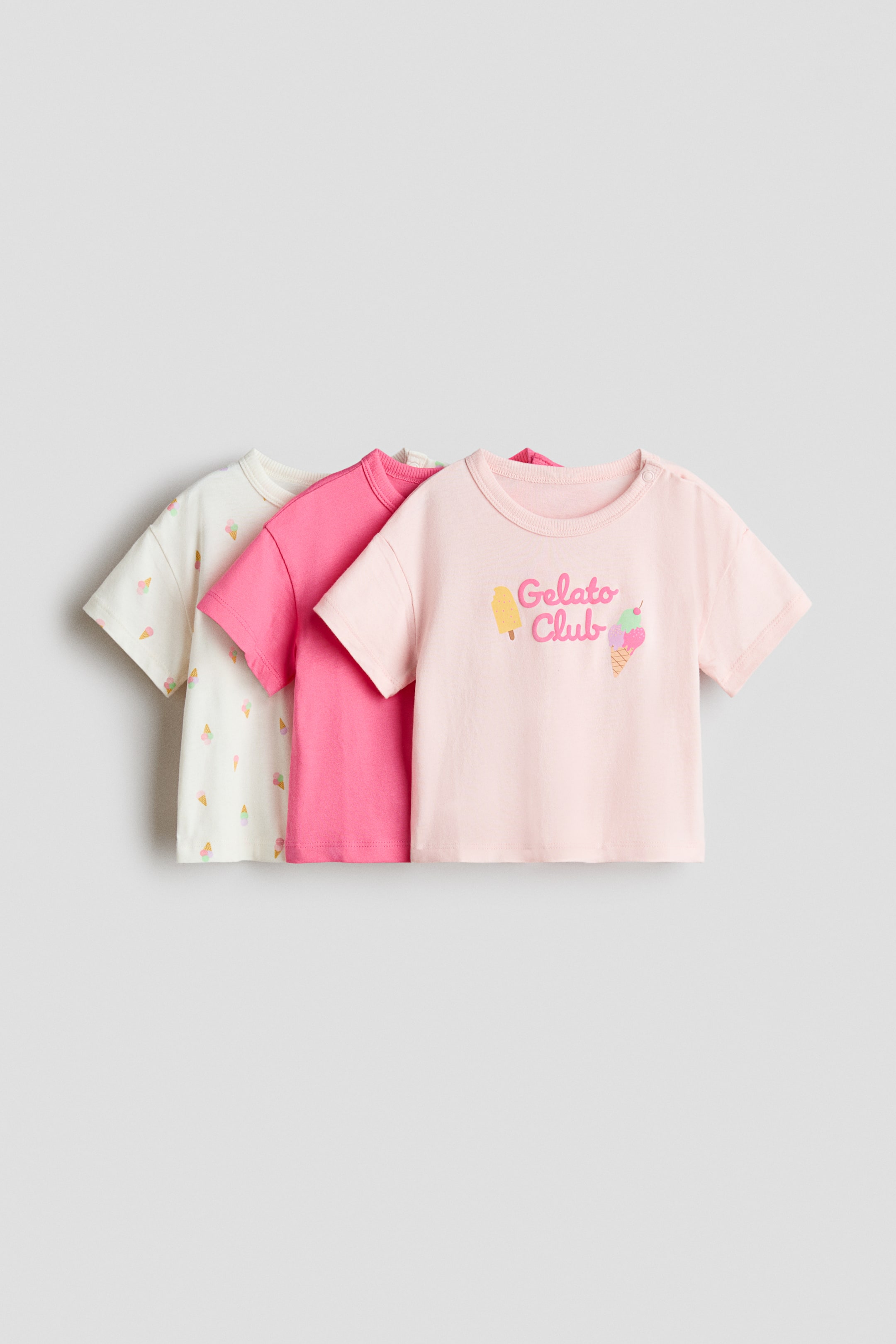 拡大画像を表示: コットンTシャツの3枚セットが展示されています。それぞれ、ショートスリーブとクルーネックのデザインです。最前面のライトピンクのシャツは正面を向いており、「Gelato Club」のグラフィックと左肩のスナップボタンが特徴です。その後ろには、ソリッドなホットピンクのシャツと、全面にアイスクリームコーンのプリントが施されたオフホワイトのシャツが重ねられています。