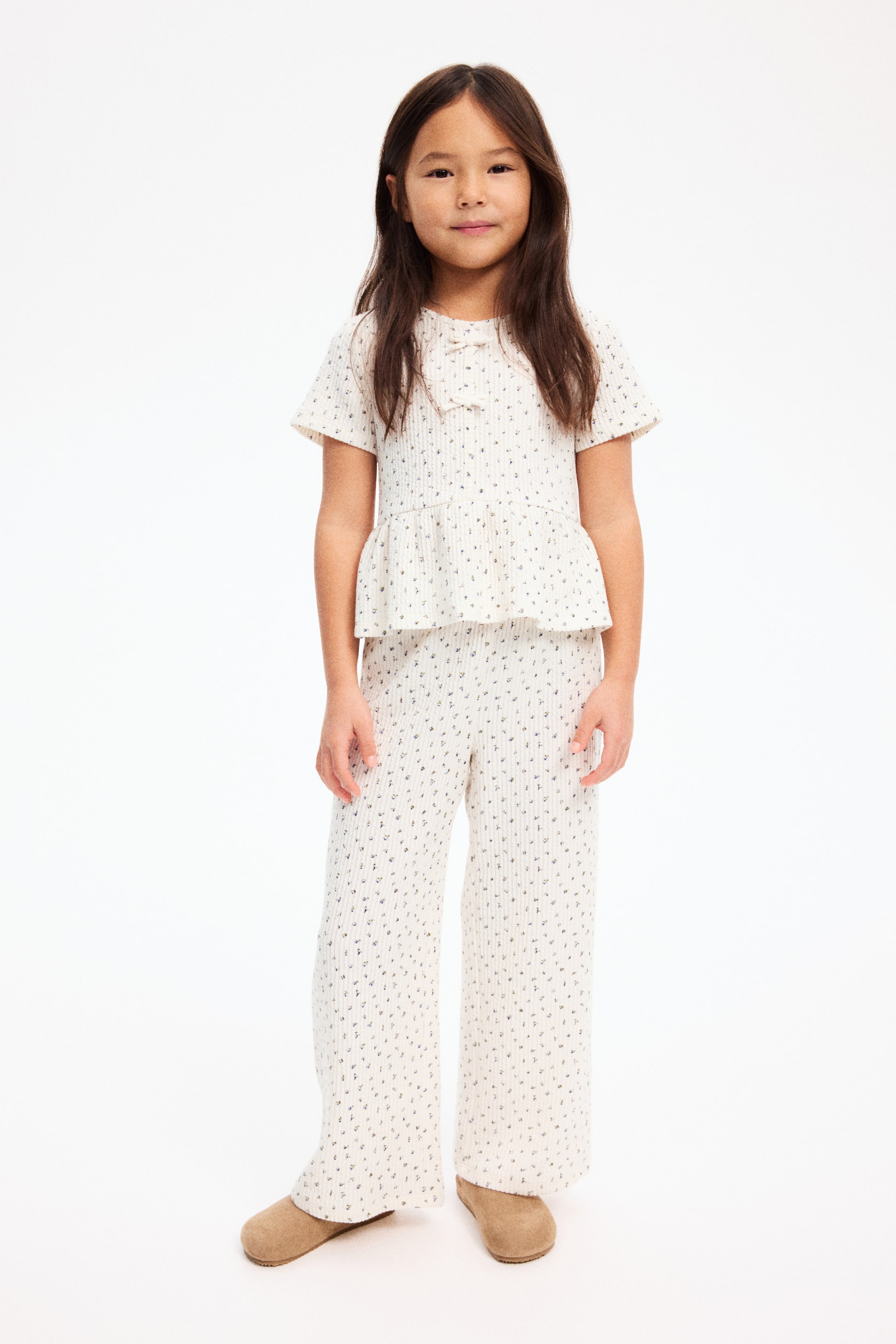 Ver imagen más grande: Un conjunto acanalado de dos piezas en blanco roto que incluye un top de manga corta y pantalones de pernera ancha, presentado de frente. El top está diseñado con escote redondo, un pequeño lazo en la parte delantera y un bajo peplum, mientras que ambas piezas presentan un sutil patrón de motas oscuras dispersas.