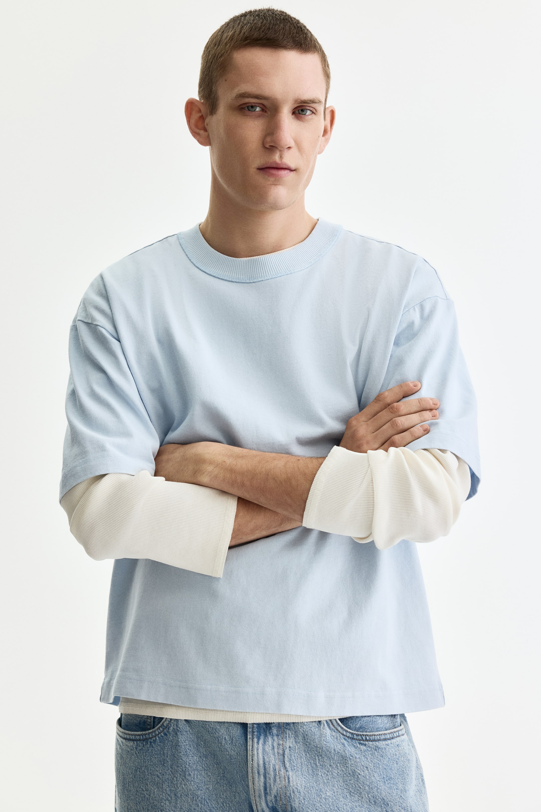 View larger image: A light blue T-shirt features a ribbed crew neck and short sleeves, displaying a relaxed, boxy fit. It is layered over a cream-colored long-sleeve shirt, with the ribbed long sleeves and hem visible underneath.