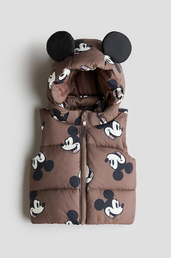 The brown puffer vest is displaying front-facing, and it is featuring an allover Mickey Mouse head print. An attached padded hood is sitting on top, which is designed with two round black Mickey Mouse ears, and a full-length brown zipper is running down the center front. - 1