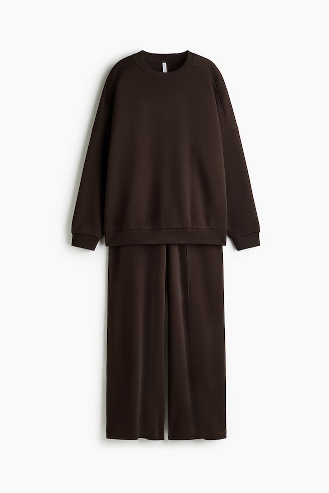 Ett mÃ¶rkbrunt tvÃ¥delat loungewear-set hÃ¤nger framifrÃ¥n, med en oversized sweatshirt med rund hals, sÃ¤nkta axlar, ribbade muddar och ribbad nederkant. De matchande byxorna med vida ben hÃ¤nger undertill och visar vertikala mittsÃ¶mmar som lÃ¶per lÃ¤ngs varje ben. - 1