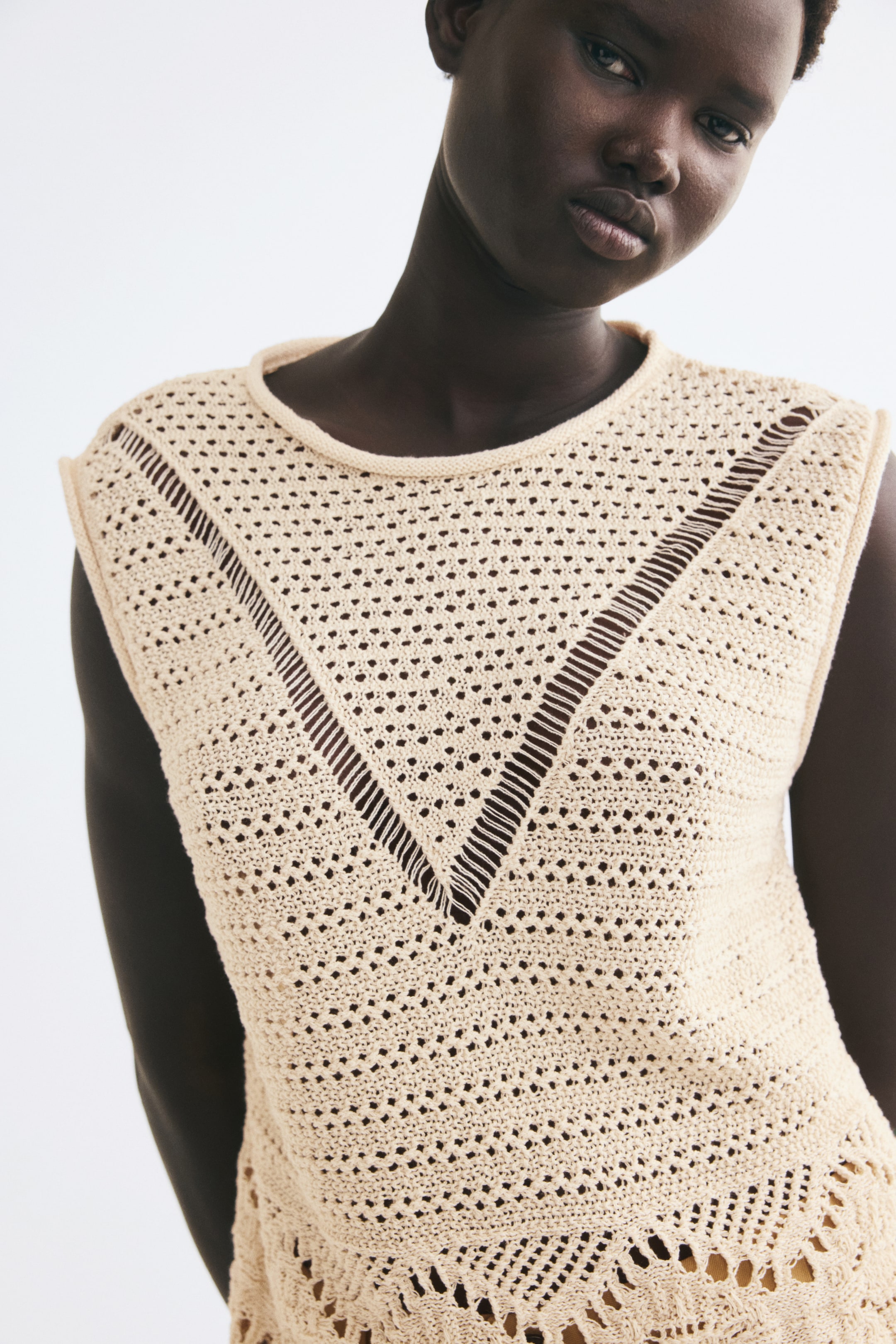 Visualizza immagine più grande: La modella indossa un top color crema senza maniche in maglia effetto crochet. Il top presenta uno scollo rotondo con bordi arrotolati e un distintivo motivo a punti aperti a forma di V sul petto.