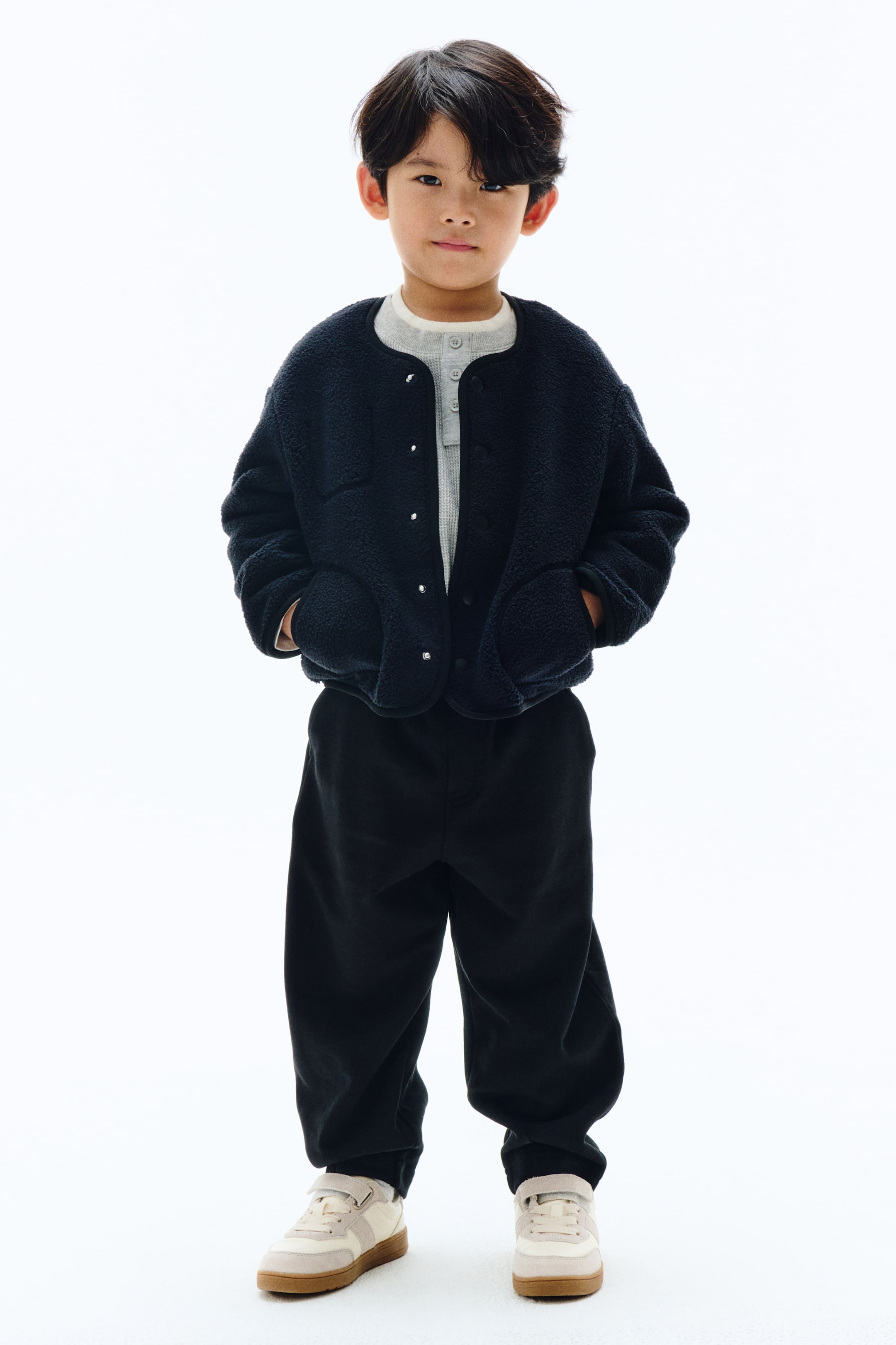 拡大画像を表示: その製品は子供用のフルアウトフィットで、スナップボタンとフロントパッチポケットが付いたダークネイビーのシェルパフリースジャケットを、ライトグレーの長袖ヘンリーネックシャツの上に着ています。ブラックのリラックスフィットのパンツがそのルックを完成させ、ベージュとオフホワイトのベルクロ付きロートップスニーカーと組み合わせています。