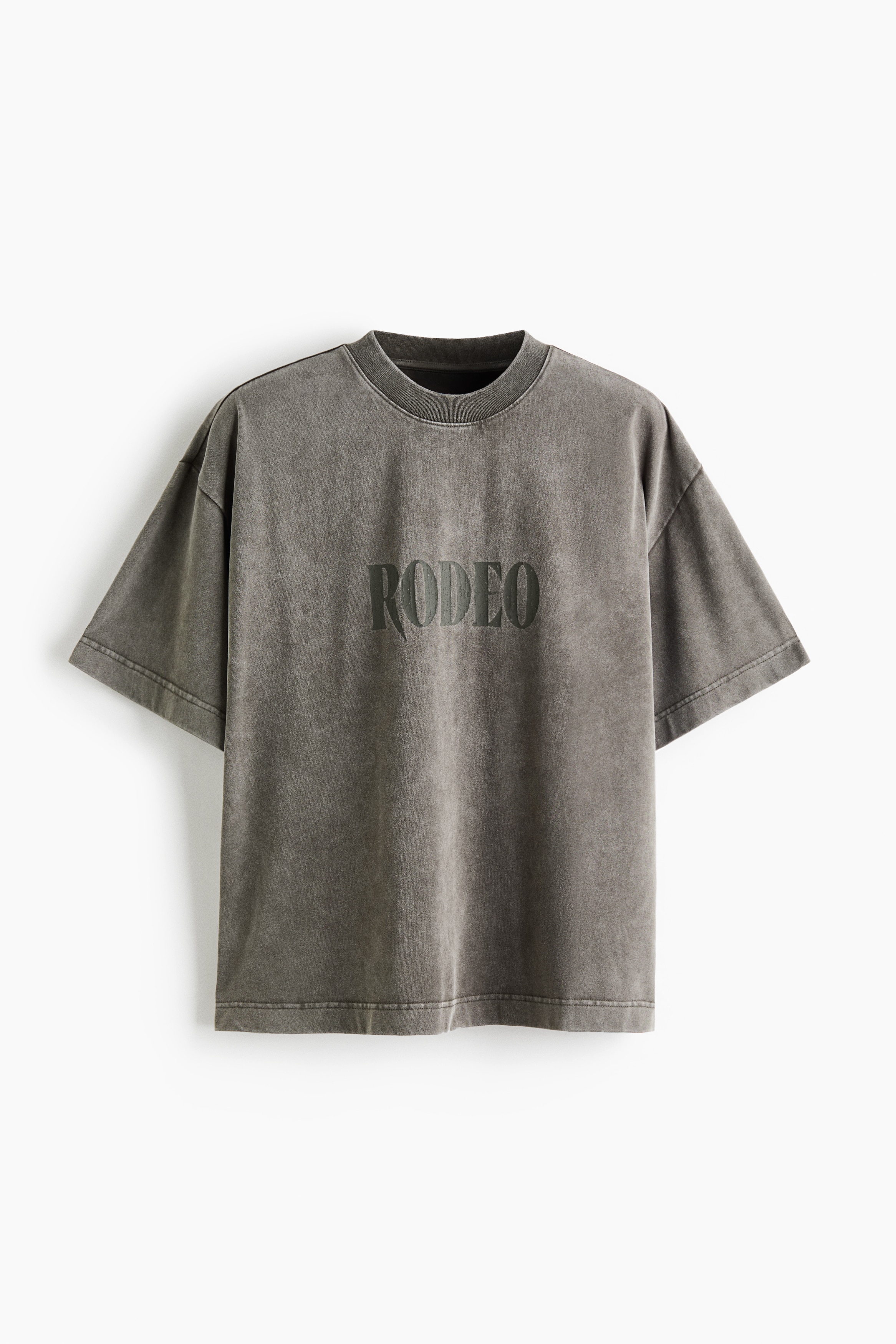An oversized, faded grey T-shirt in heavyweight cotton jersey is being worn by the model, featuring a printed "RODEO" graphic across the chest. The T-shirt has dropped shoulders and a ribbed crew neck, styled with beige trousers and a red-and-black plaid flannel shirt tied around the waist.
