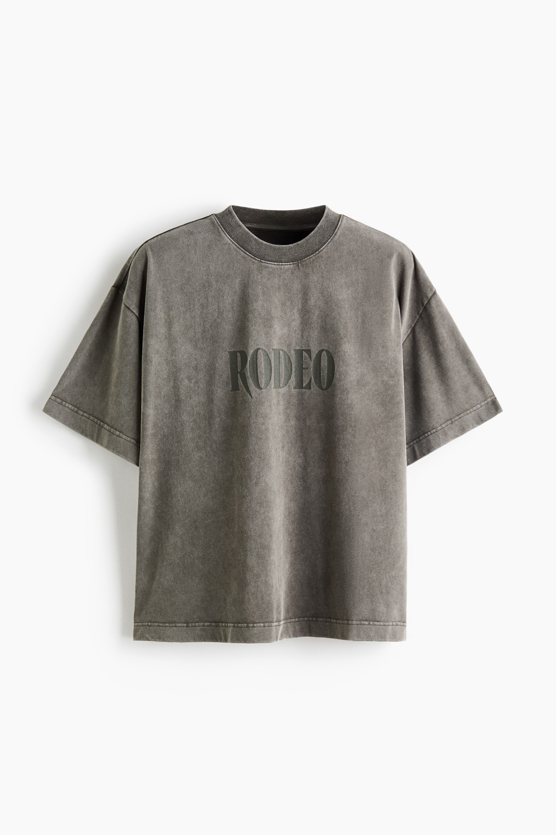 View larger image: A faded grey, acid-washed oversized T-shirt is shown from the front, featuring a ribbed crew neckline, dropped shoulders, and wide short sleeves. The word "RODEO" is printed across the centre chest in a dark, stylized font.