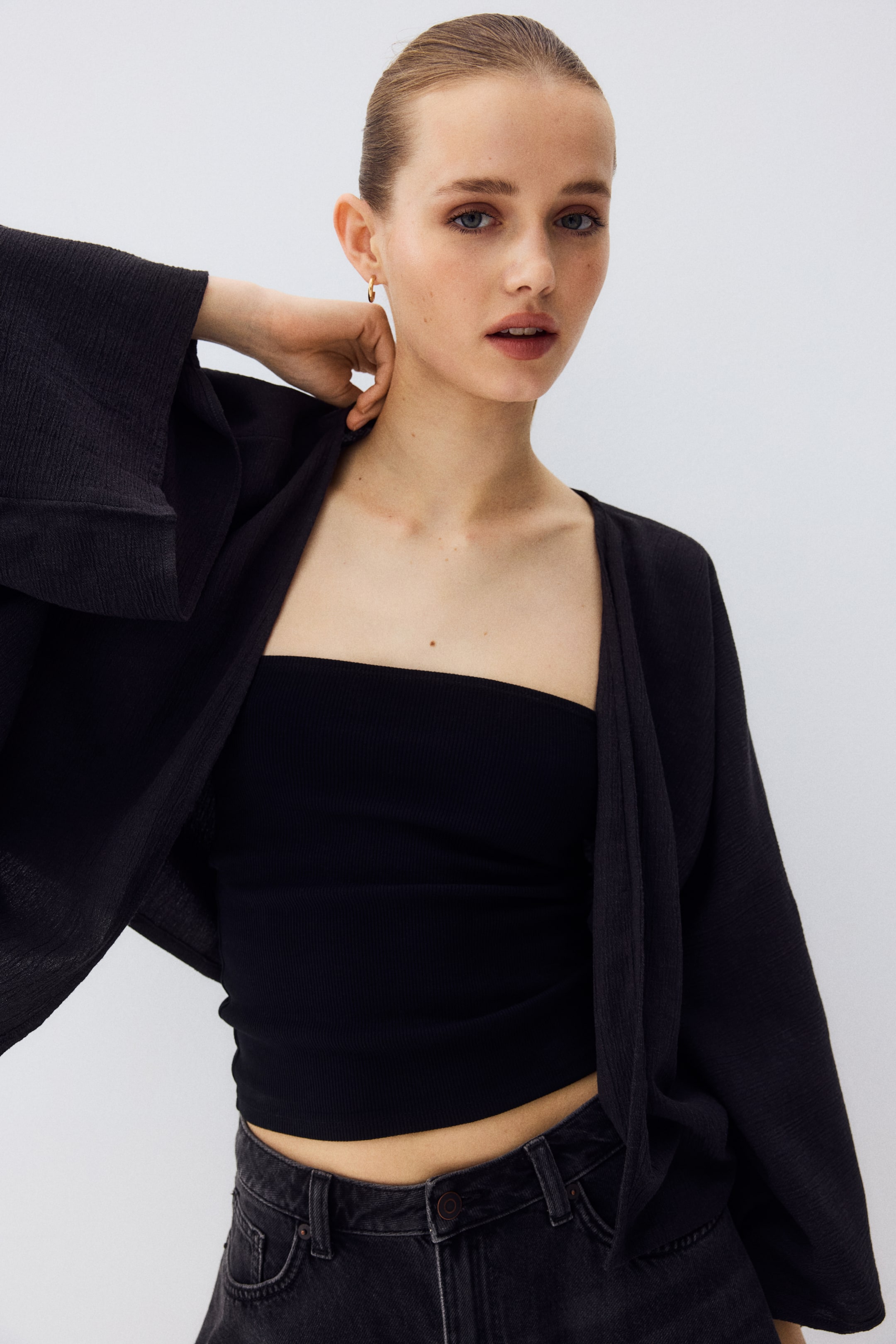 View larger image: A black open-front cardigan is showcased from the front, highlighting its relaxed drape over a black tube top. It comes with wide, flowing sleeves and a crinkled texture.