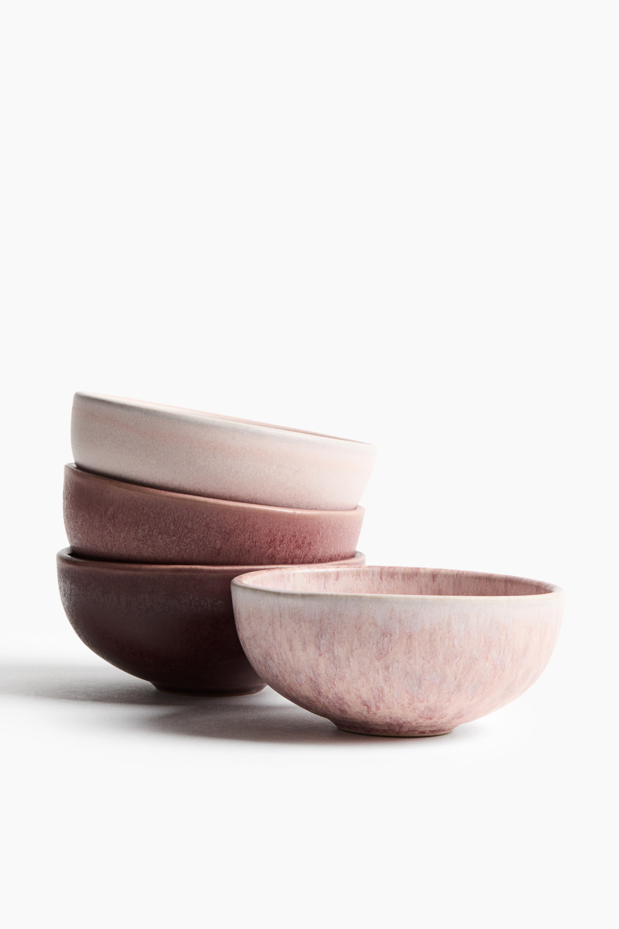 View larger image: Four stoneware bowls display a reactive glaze in varying shades of pink, mauve, and deep brown, with three of the bowls stacked and one bowl positioned to the right. The bowls feature a classic rounded shape with a footed base and a textured, variegated surface finish.
