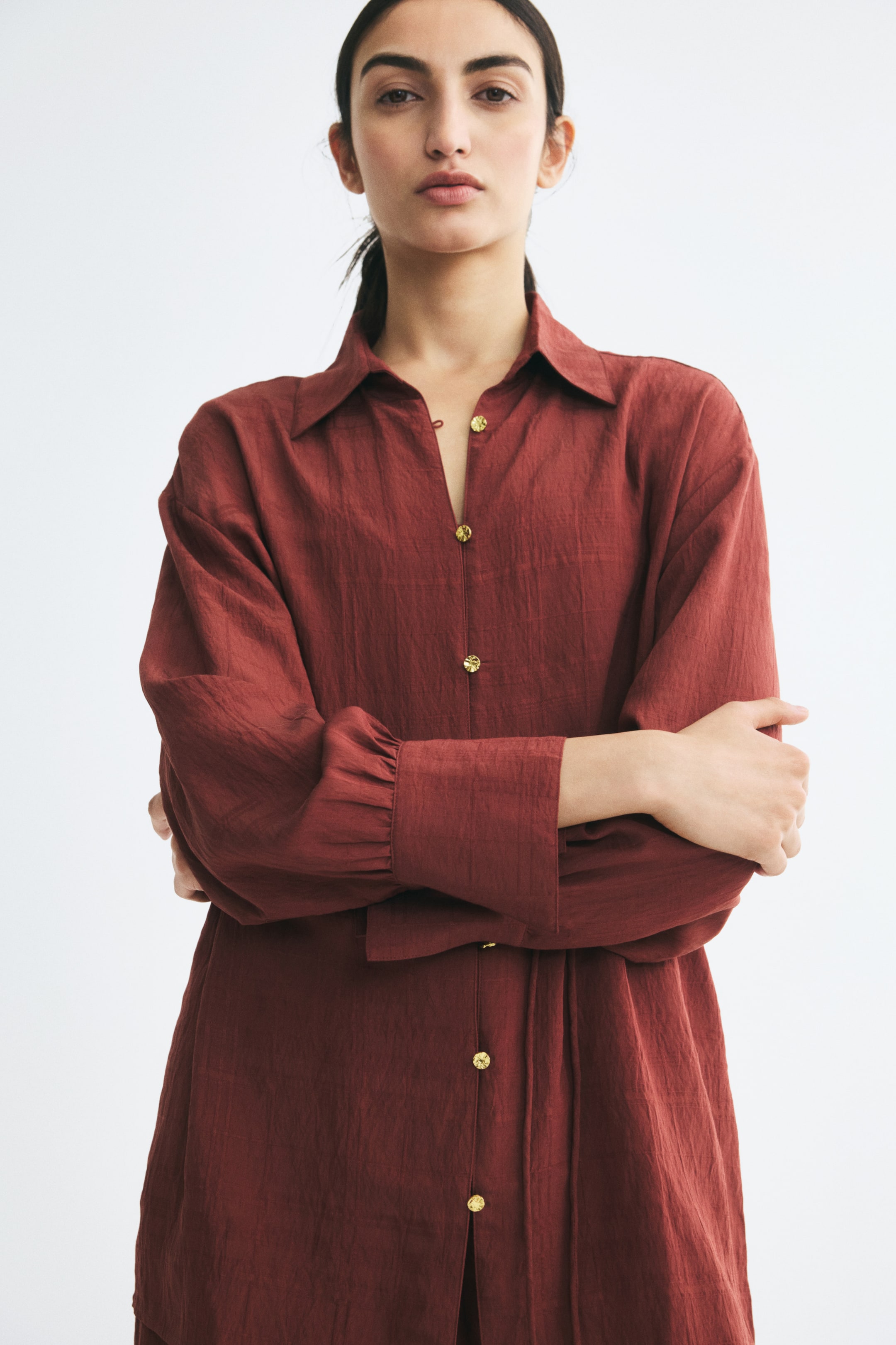 View larger image: A rust-coloured, long-sleeved shirt is being worn by the model, who is posing with crossed arms. The shirt features a pointed collar, a partially unbuttoned front with textured gold-tone buttons, and wide cuffs with gathered detailing.