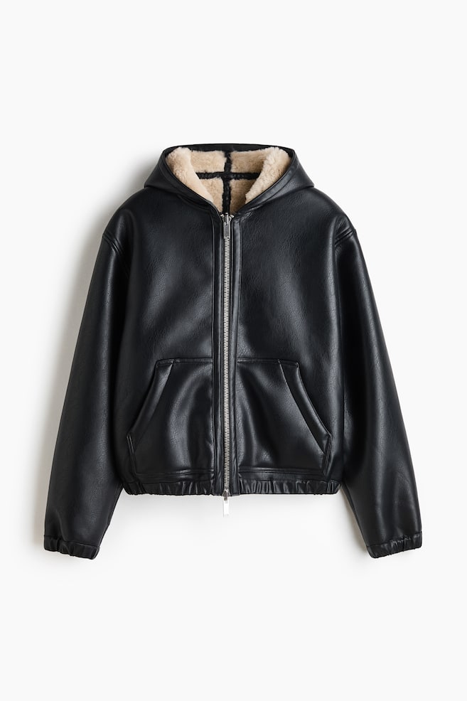 A black faux-leather hooded jacket is shown from the front, featuring a silver-tone zipper closure and a plush beige shearling lining visible inside the hood. The jacket is displaying two front slip pockets, an elasticated hem, and elasticated cuffs. - 1