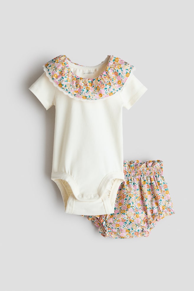 Un body à manches courtes couleur crème et un bloomer à imprimé floral sont présentés en ensemble, vus de face. Le body présente un col volanté floral avec une délicate bordure en dentelle, et des boutons-pression sont visibles à l'entrejambe. Le bloomer affiche une taille élastiquée recouverte et des volants à la taille et autour des jambes. - 1