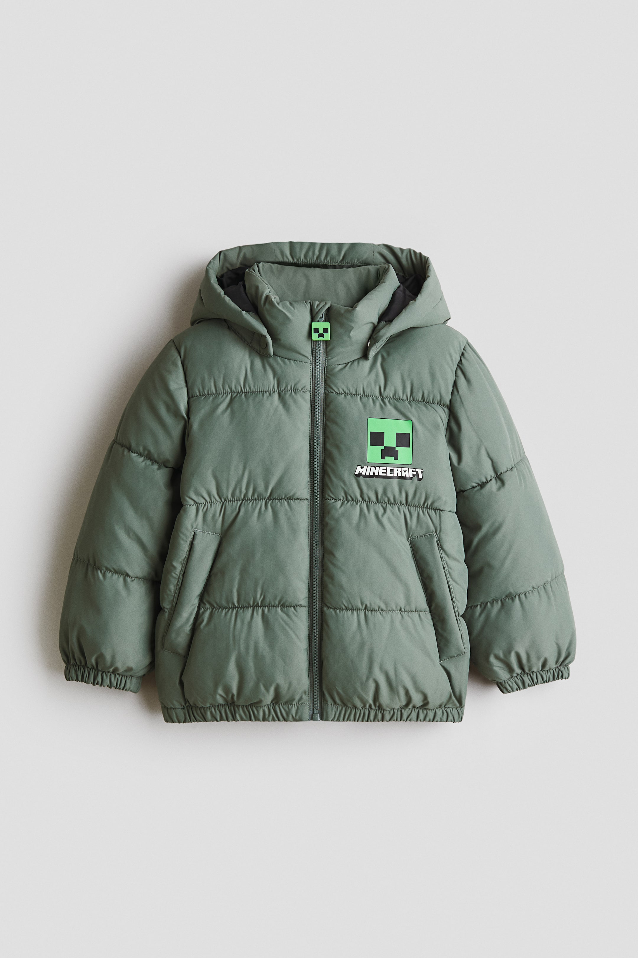 View larger image: Water-repellent puffer jacket is presented front-facing, featuring a sage green colour with horizontal quilting and an attached hood. A full-length zip with a green pixelated face zip pull is closing the front, while a matching green pixelated face patch with 'MINECRAFT' text is appearing on the left chest, complemented by side pockets and elasticated cuffs and hem.