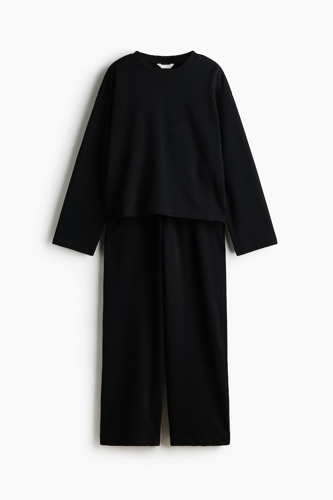 A black two-piece loungewear set is presenting front-facing, featuring a loose, long-sleeved crew neck top and wide-leg pants. The cropped top is resting over the full-length pants, with both pieces displaying a smooth, dark fabric. - 1