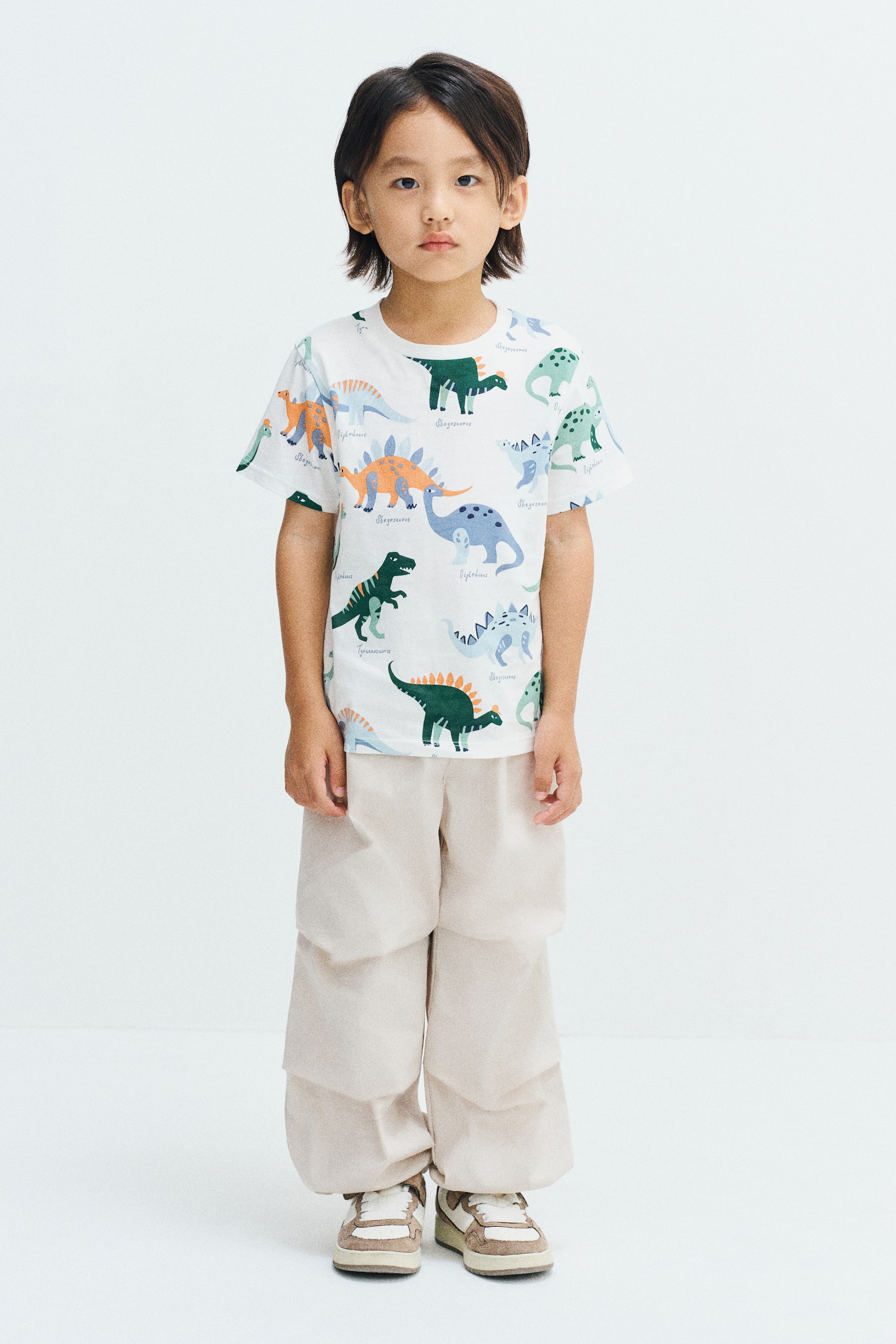 View larger image: The model is wearing a white short-sleeved tee featuring a colourful all-over dinosaur print and a rib-trimmed round neckline. The tee is styled with loose, light beige cargo trousers and sneakers.