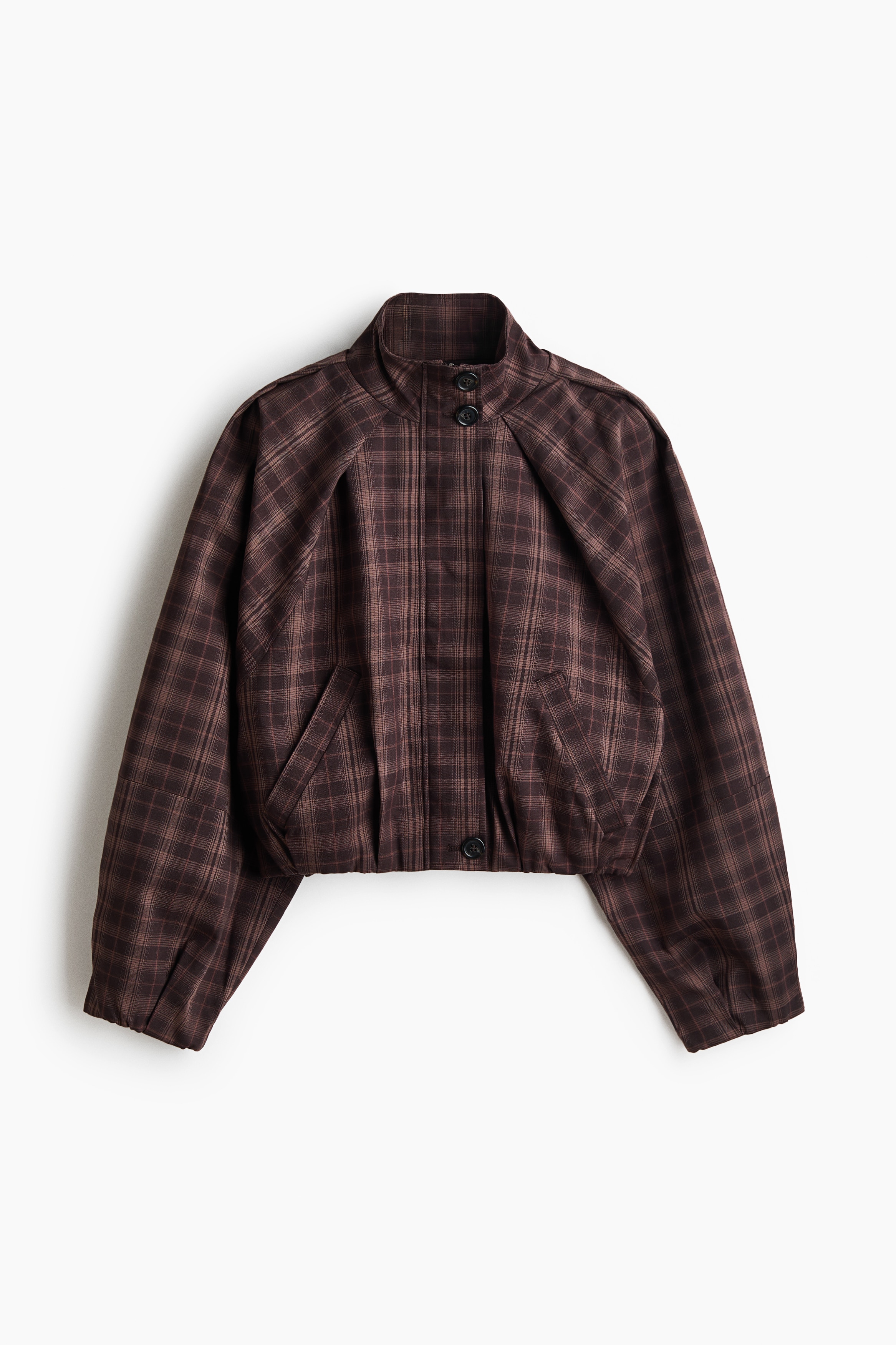 La modelo lleva puesta una chaqueta corta a cuadros marrón, con cuello mao, tapeta cortaviento con botones y mangas raglán. La chaqueta cropped se lleva sobre una prenda superior de color claro y se combina con una minifalda a cuadros del mismo estampado.